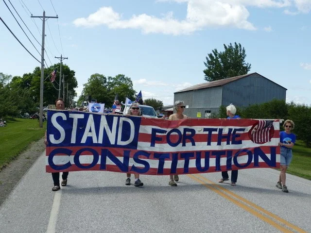 Group of people walking on a rural road holding a large banner that says "Stand for the Constitution," with American flags and blue-themed decorations, under a partly cloudy sky.