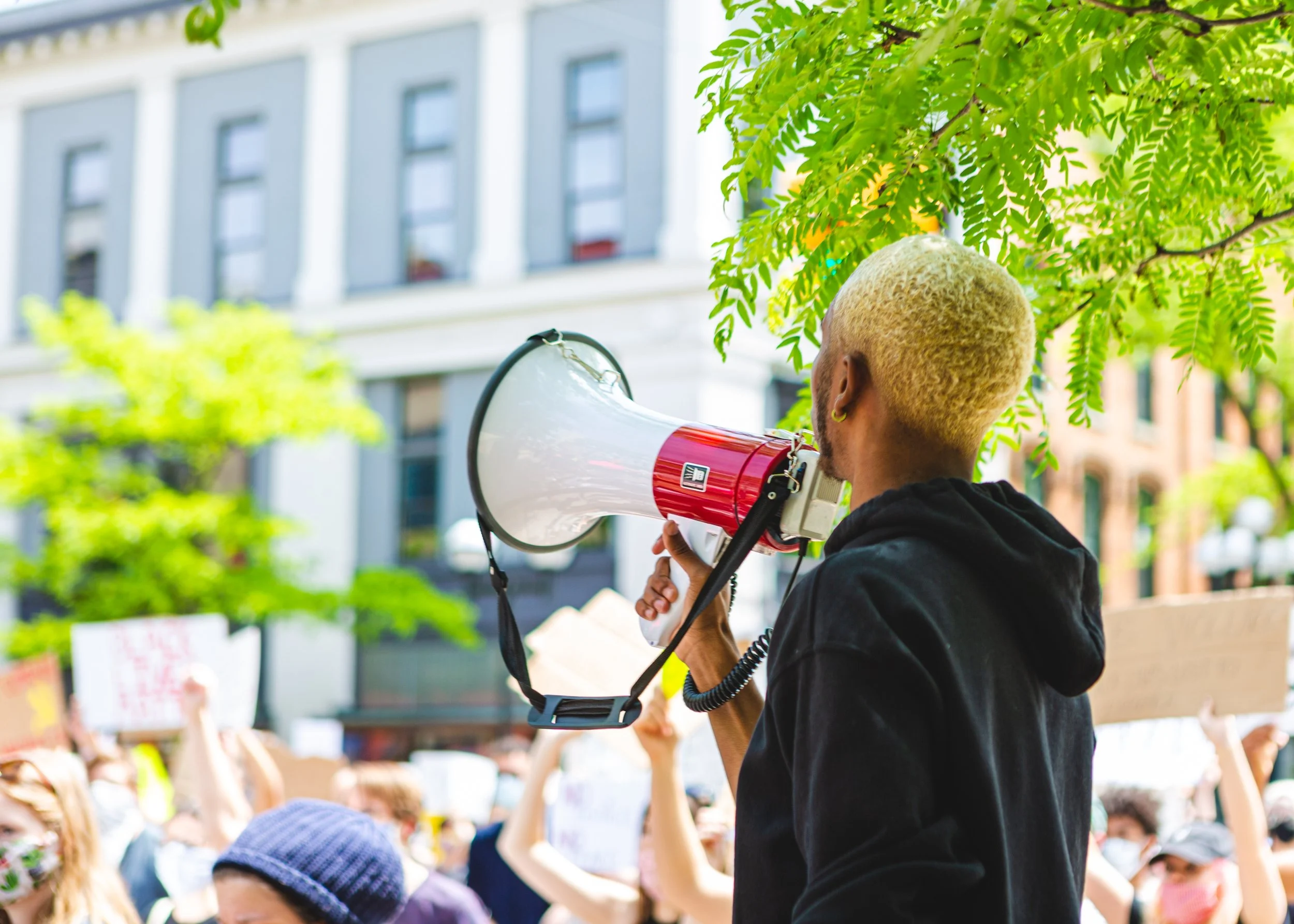Person with short blonde hair using a megaphone at a protest or rally, with a crowd holding signs and wearing masks.