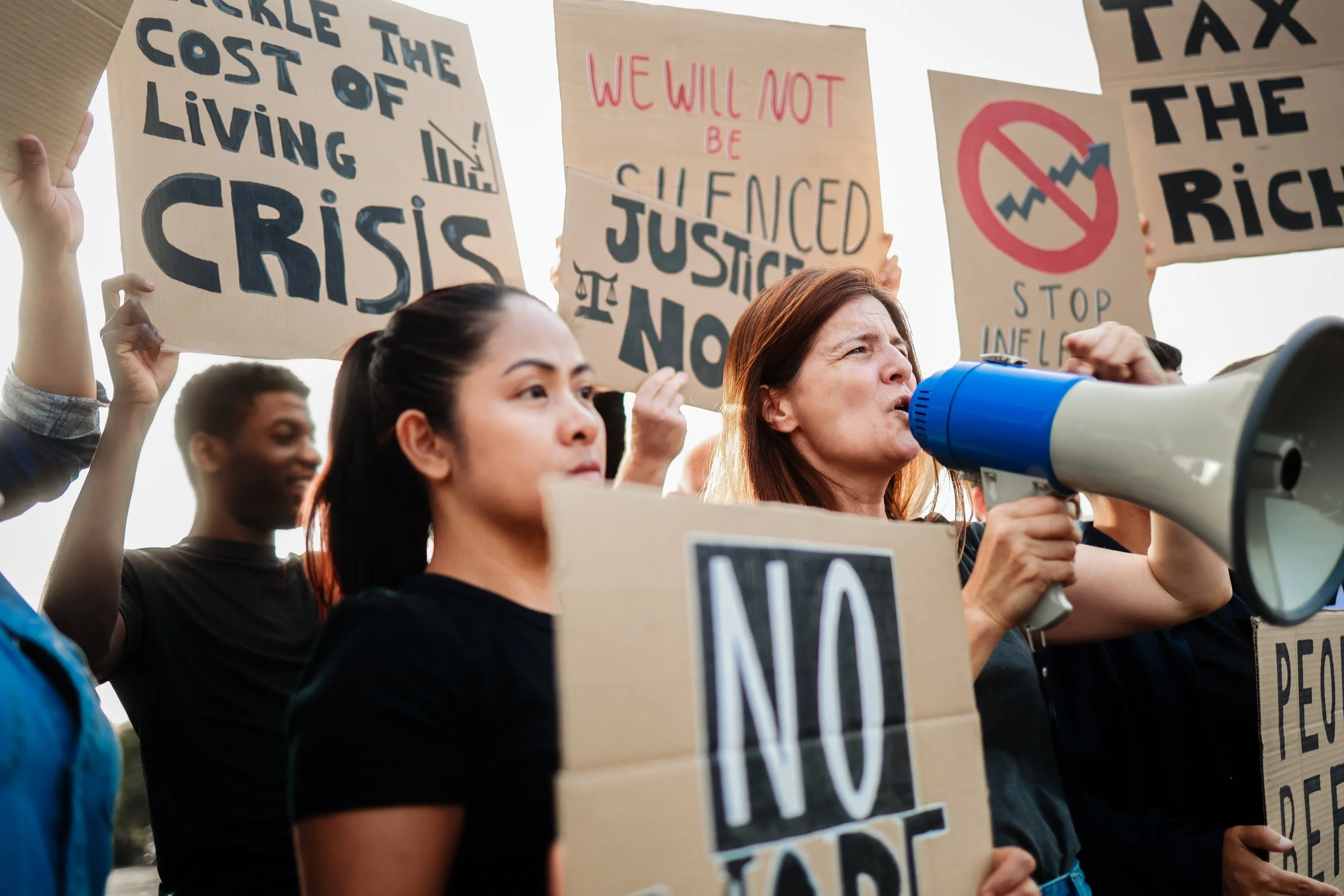 Protesters holding signs that demand action on economic issues, including stopping inflation, resisting the cost of living crisis, and advocating for justice and fair wages, with a woman speaking into a megaphone.