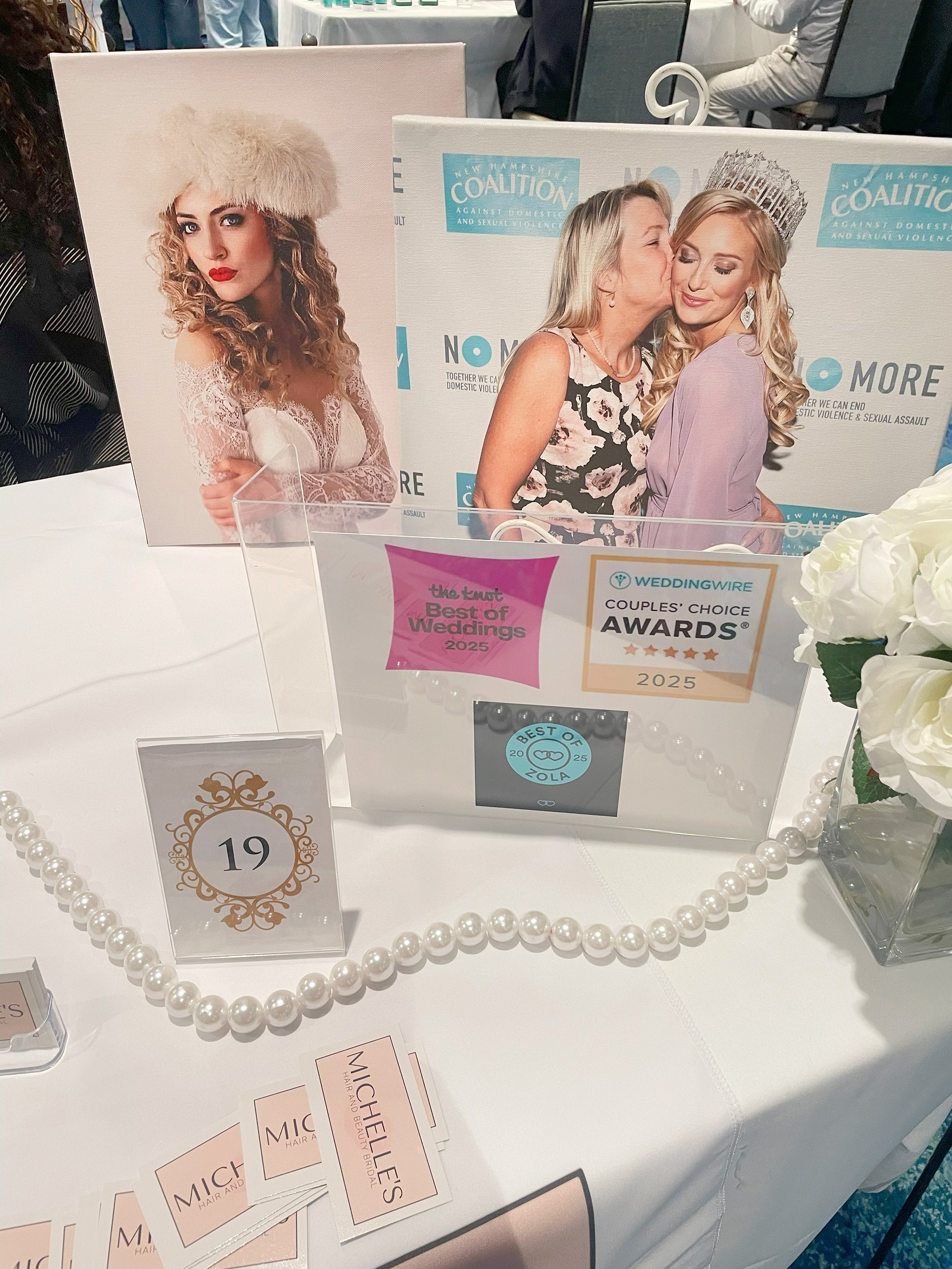 Table display at an awards event with photos of women, award logos, a pearl necklace, and business cards.