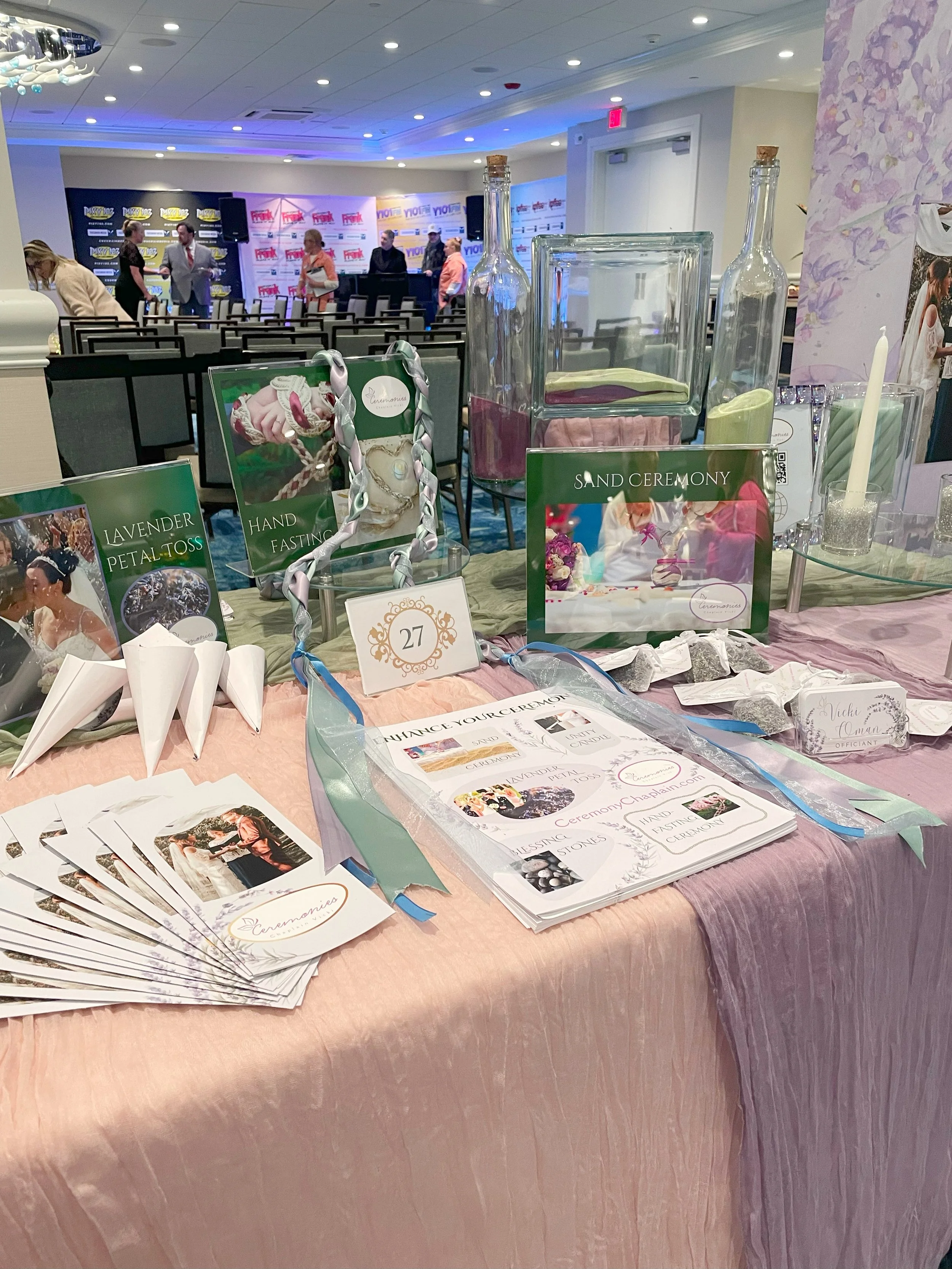 Sweet table setup at a wedding expo, displaying sand ceremony materials, brochures, and promotional items with chairs and a stage in the background.
