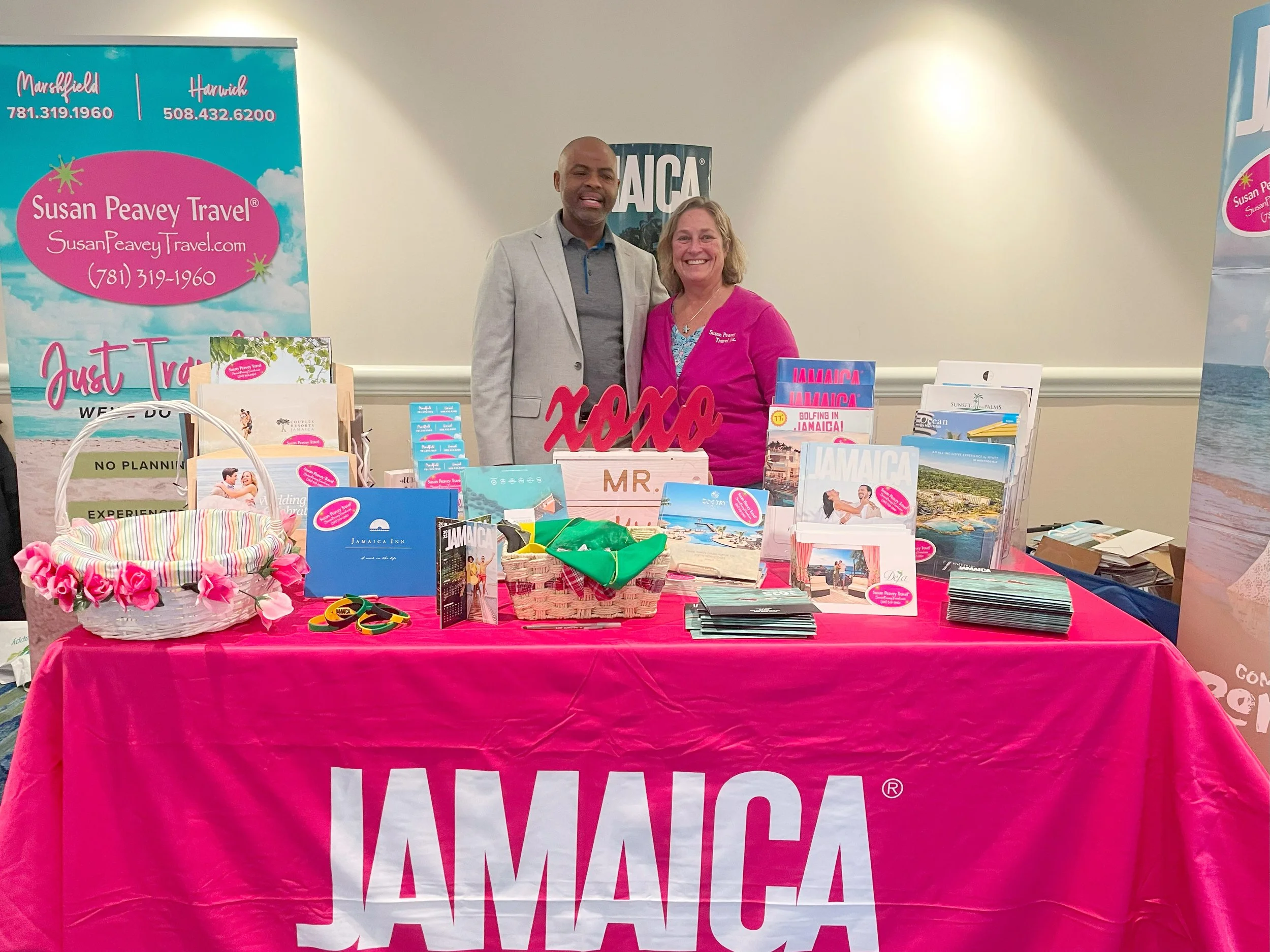 Two people standing behind a travel booth with a pink tablecloth that has 