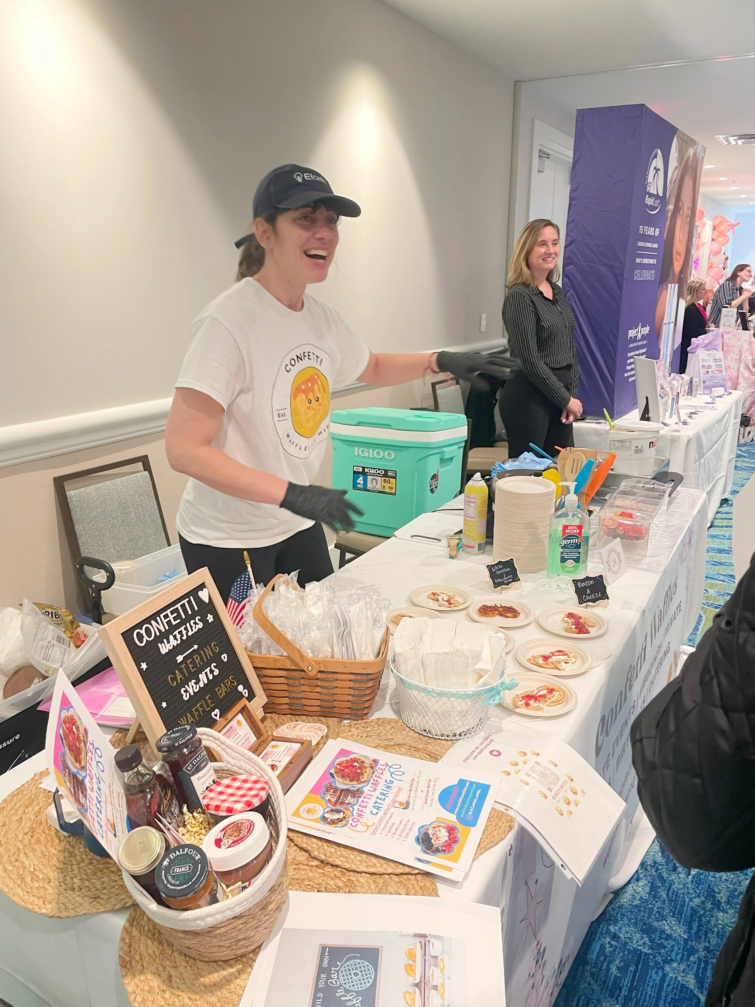 A woman at a dessert booth, smiling and gesturing, with plates of mini desserts, condiments, and promotional materials on the table.