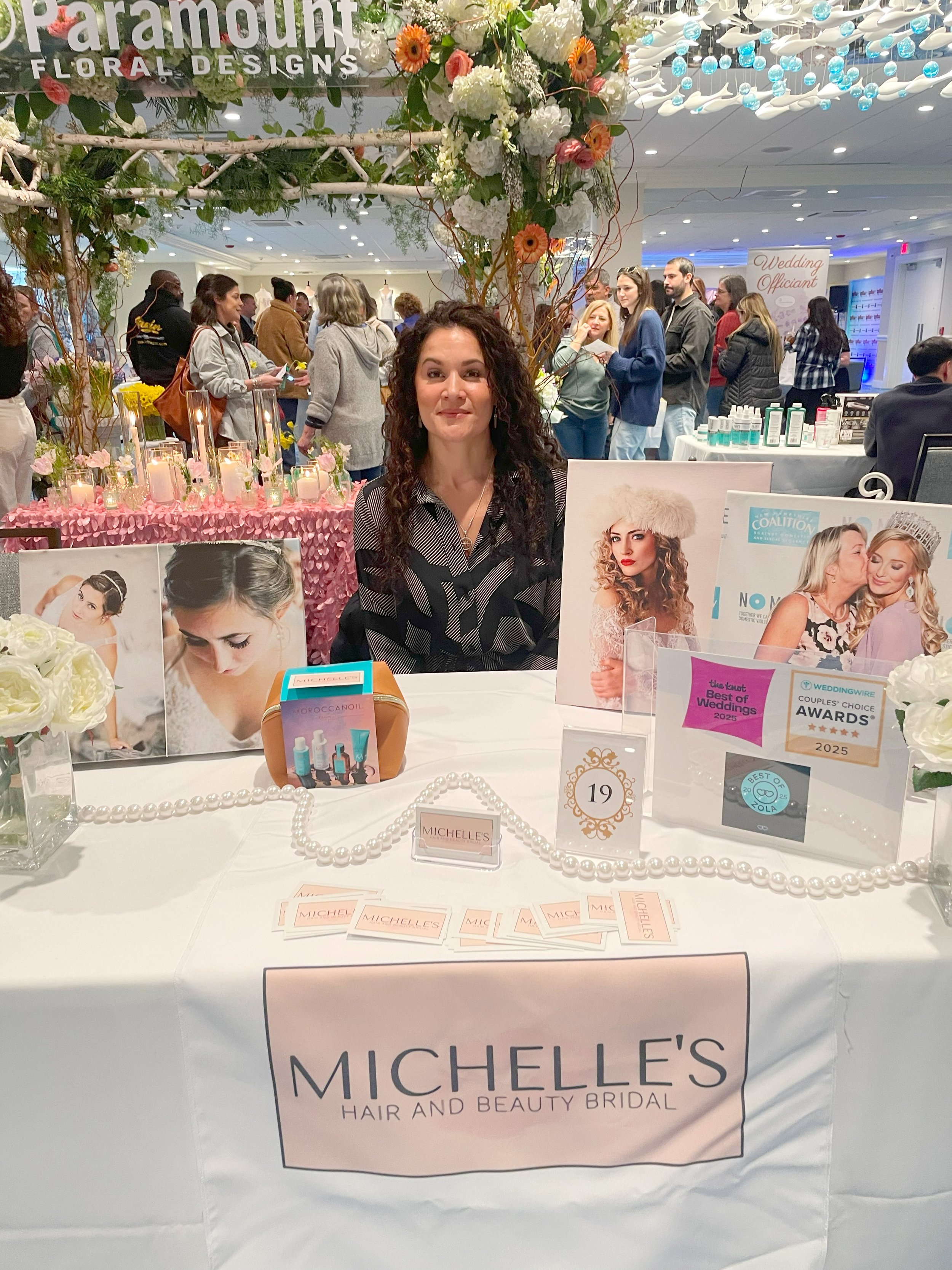 Woman sitting at a booth promoting Michelle's Hair and Beauty Bridal with floral decorations and photos of bridal hair and makeup. The booth displays products and awards related to wedding beauty services, set in a busy event space.
