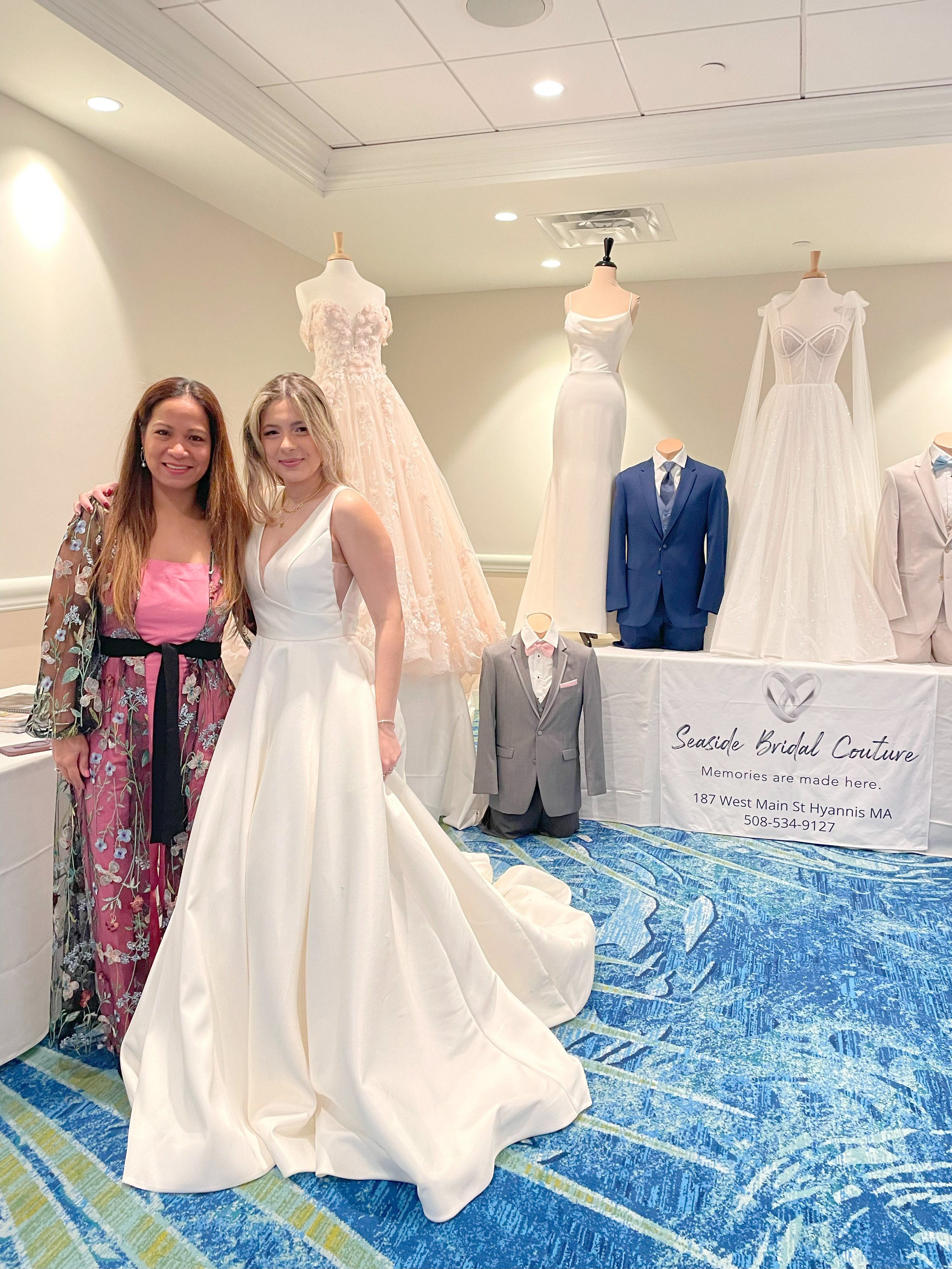 Two women standing in front of a display of wedding dresses and mannequins dressed in suits at a bridal boutique.