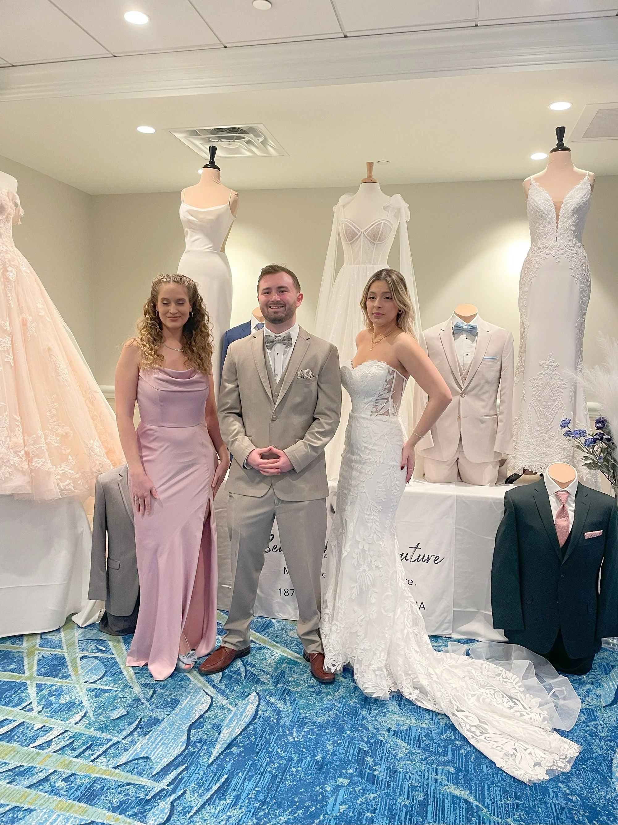 People dressed in wedding attire standing in front of mannequins wearing wedding dresses and suits at a bridal shop.