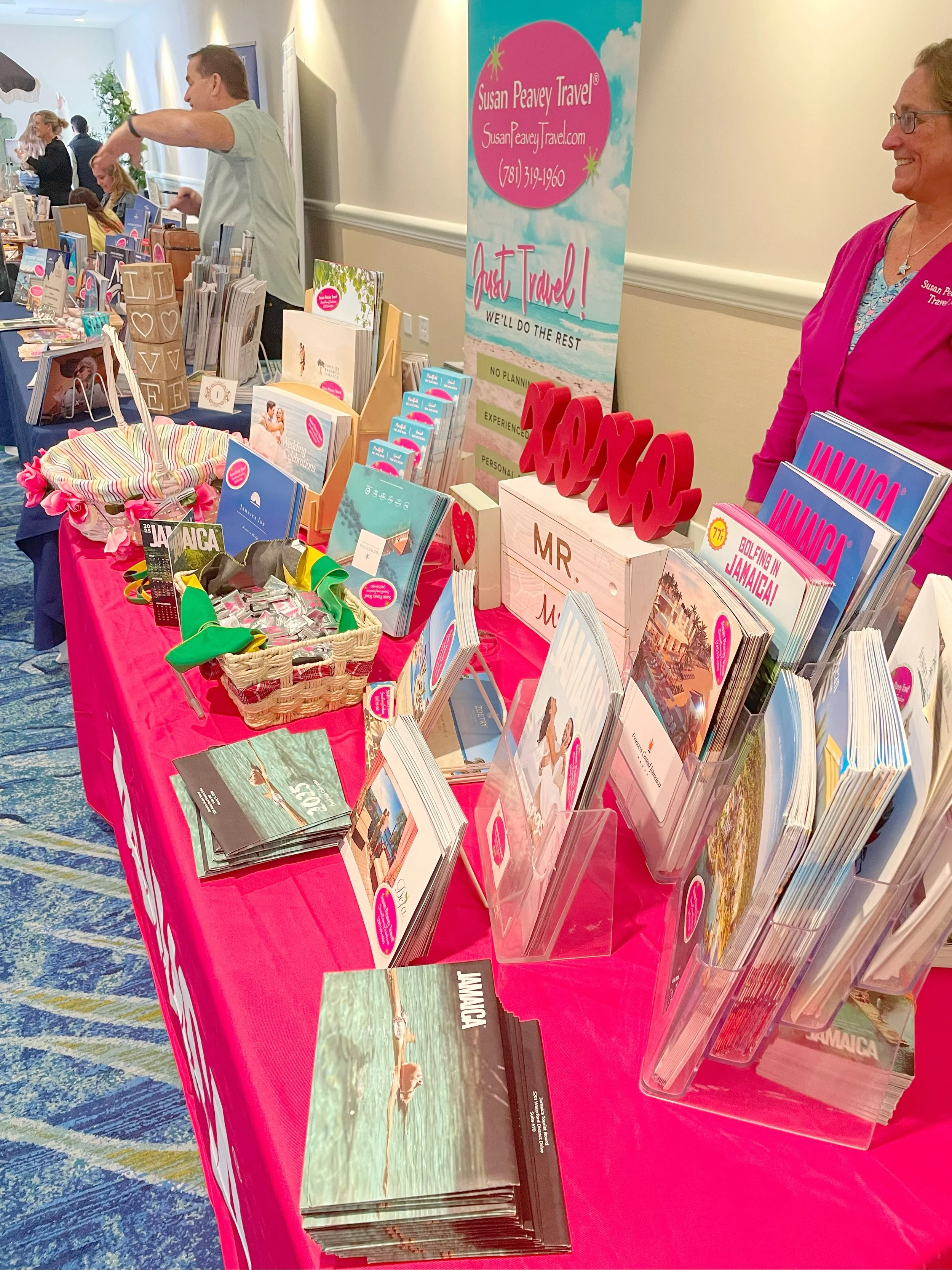 A travel agency booth with brochures, flyers, and souvenirs promoting Jamaica with the word "Jamaica" prominently displayed on materials and a large red decorative "Jamaica" sign. A woman in a pink shirt is standing behind the table, and other booths