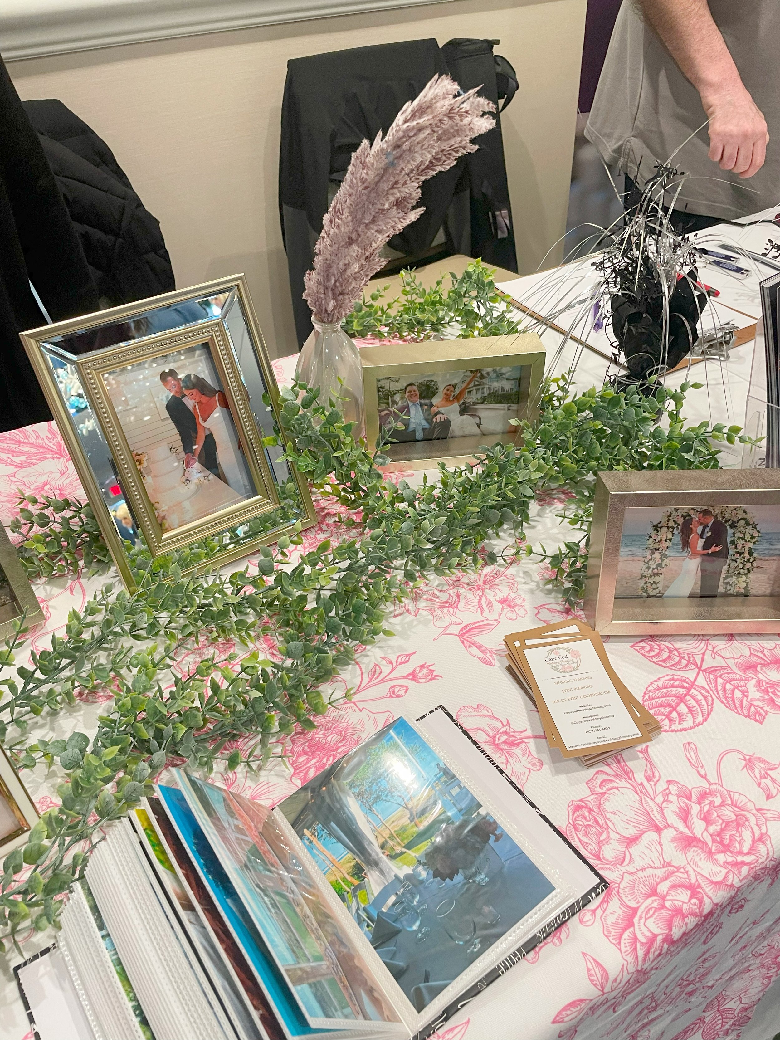 A table decorated with framed photographs of a couple, green leafy garland, a vase with tall, feathery pink flowers, brochures, and various items, likely part of a wedding or celebration display.
