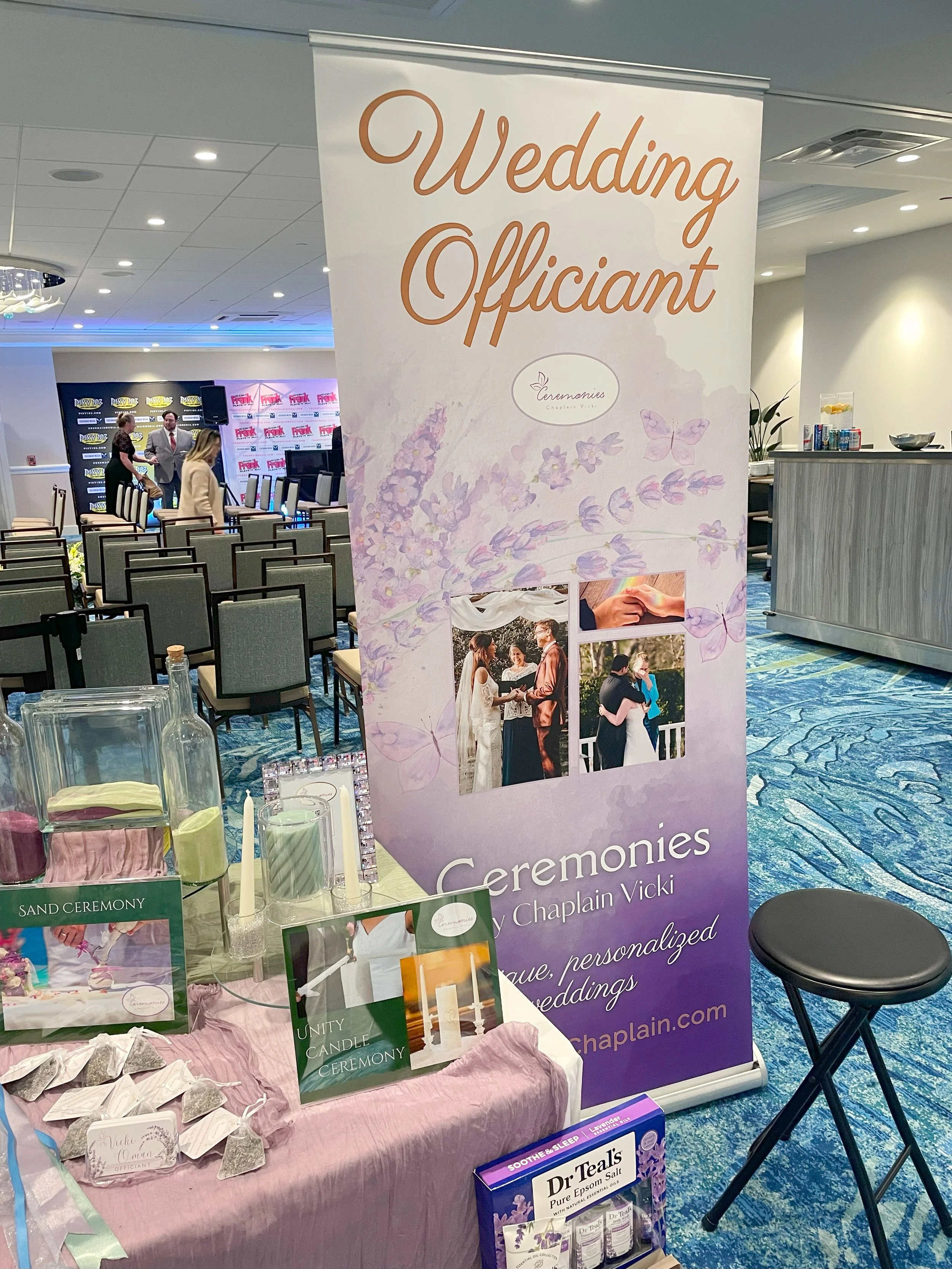 Wedding officiant booth with a tall banner displaying the text 'Wedding Officiant' and images of couples at an outdoor wedding. The booth has candles, small bags labeled 'Sand Ceremony,' and informational materials. The event appears to be in a banqu