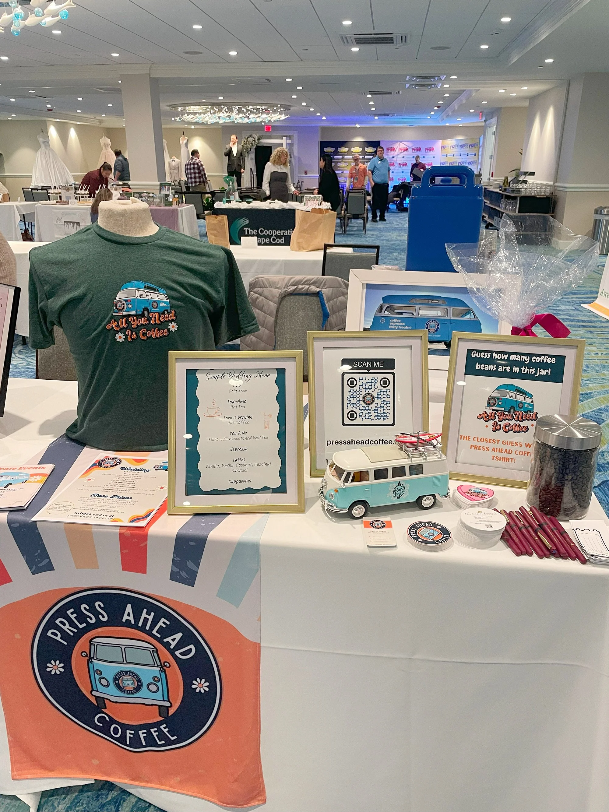 Table with sign promoting press coffee, featuring a T-shirt with the slogan 'All You Need Is Coffee,' a miniature van model, and signs asking to guess coffee beans and scan a QR code, set in a large room with people and mannequins in the background.