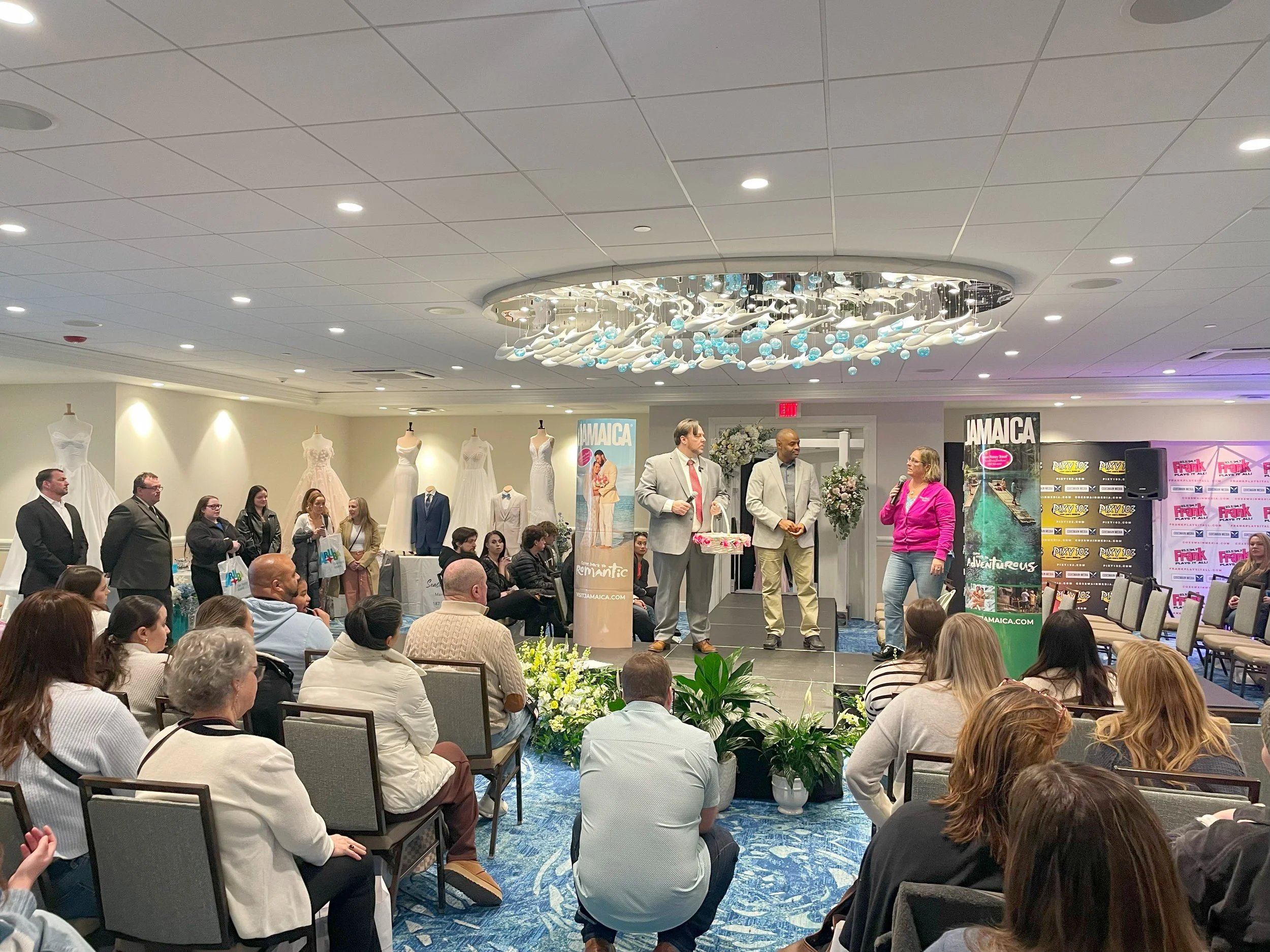 Group of people attending a presentation or event in a hotel conference room, with mannequins dressed in wedding gowns and posters advertising Jamaica