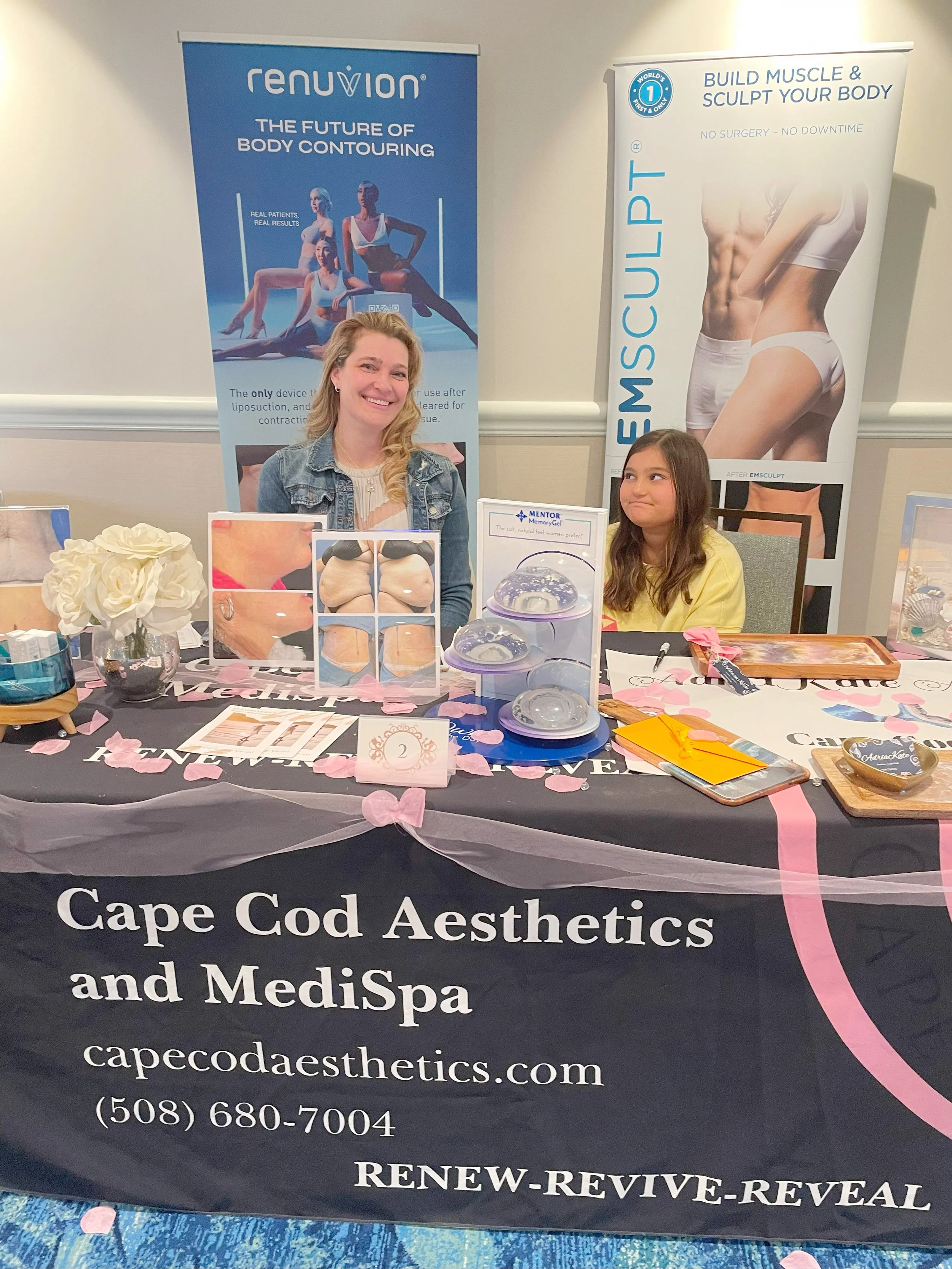 A woman and a girl sitting at a booth for Cape Cod Aesthetics and MediSpa, with promotional banners for body contouring and muscle sculpting in the background. The table is decorated with pink flower petals and various promotional materials.