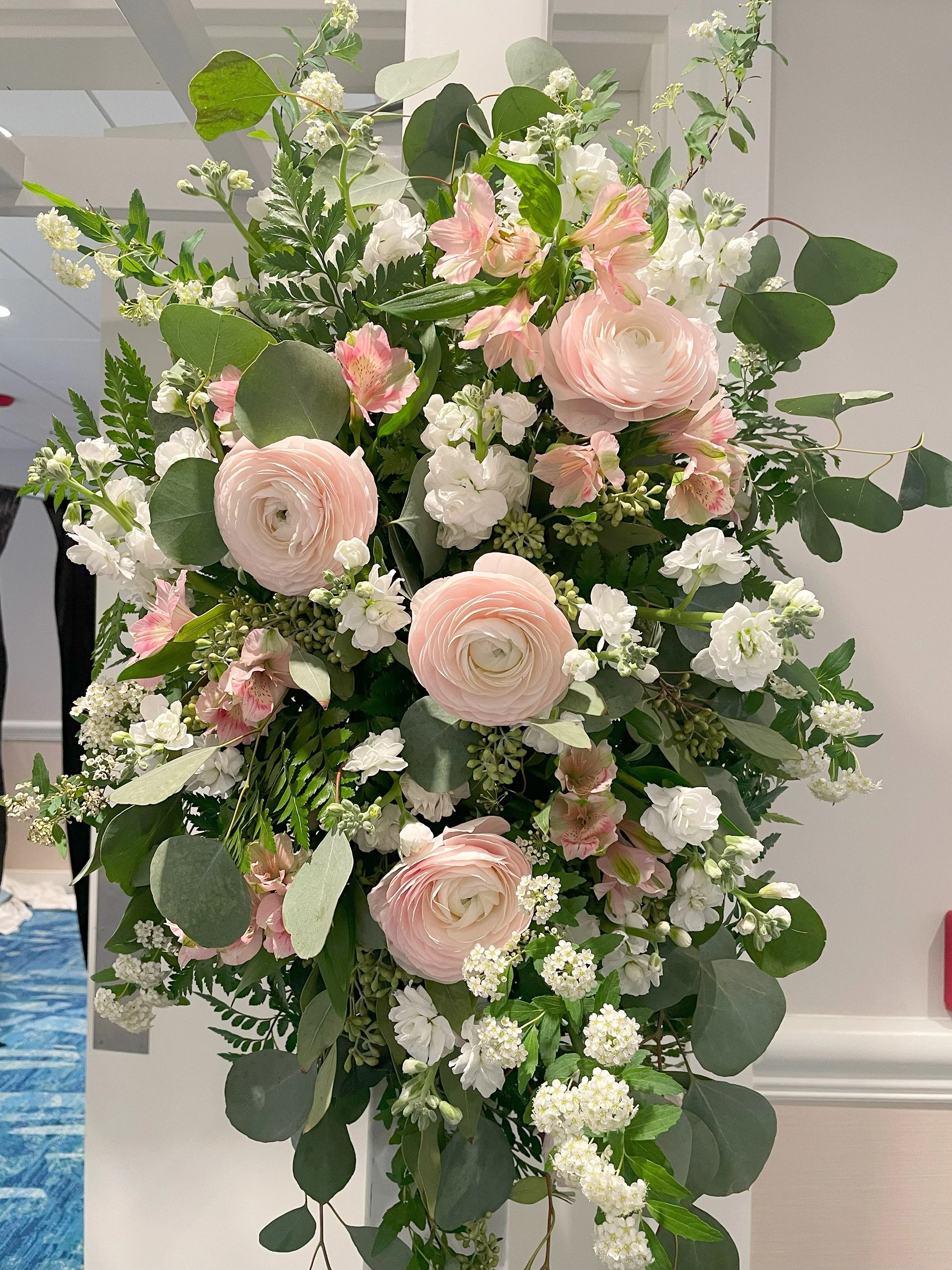 A large flower arrangement with pink and white roses, white ranunculus, pink and white alstroemeria, white stock, and various green foliage, including eucalyptus leaves and ferns.
