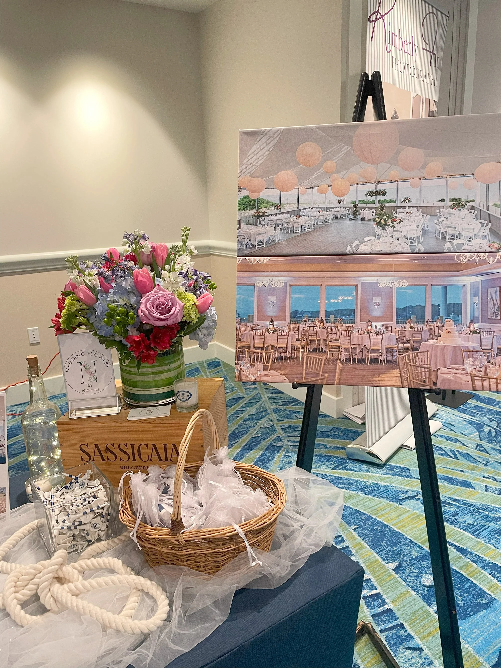 Decorative display featuring a bouquet of pink and purple flowers in a striped green vase, a wooden box labeled 'SASSICAIA,' a basket with tulle, a clear bottle, and a container with small white rolled items. Behind the display, there are large poste