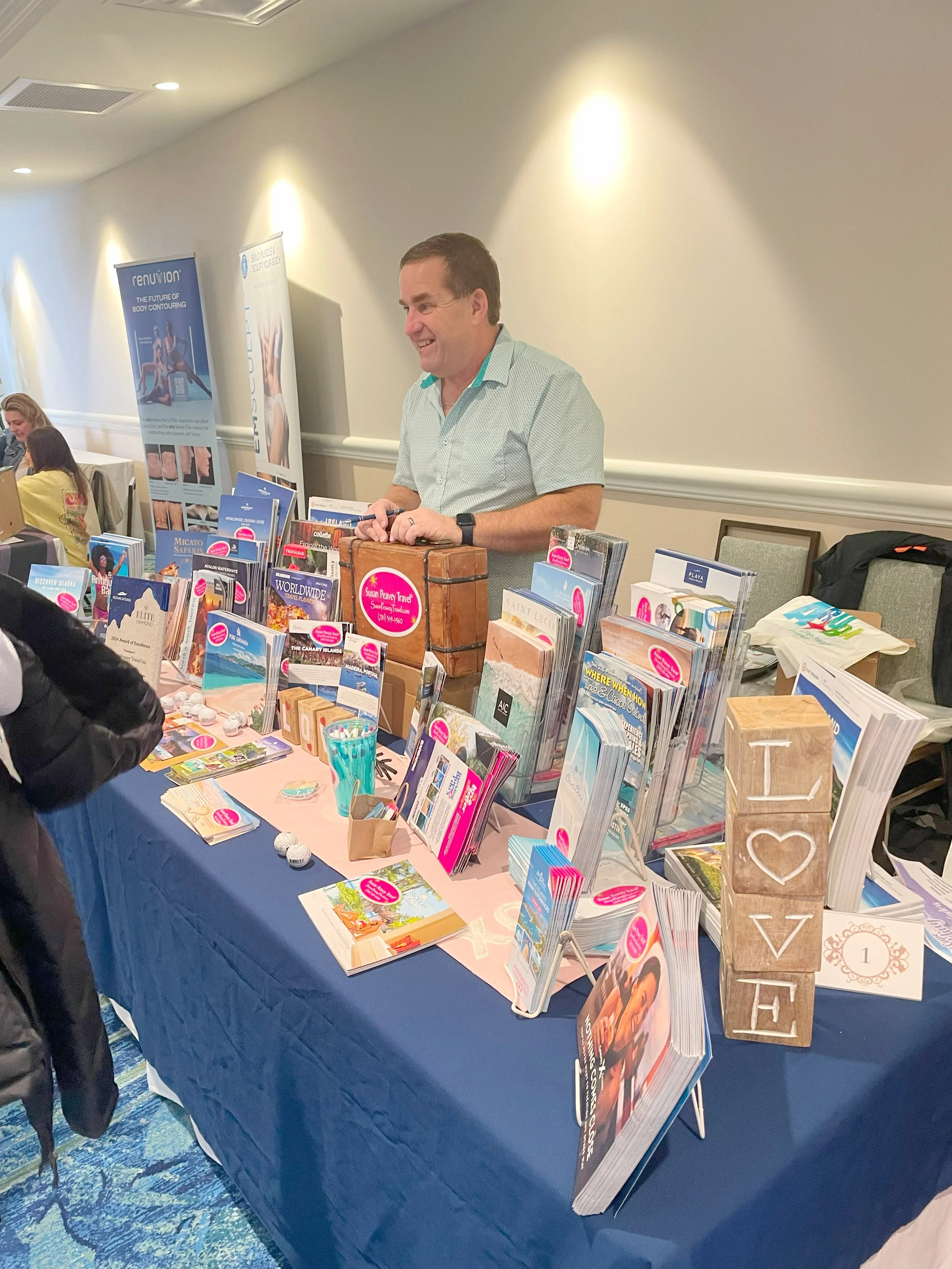 A man standing behind a table of travel brochures and books at an indoor event, smiling.