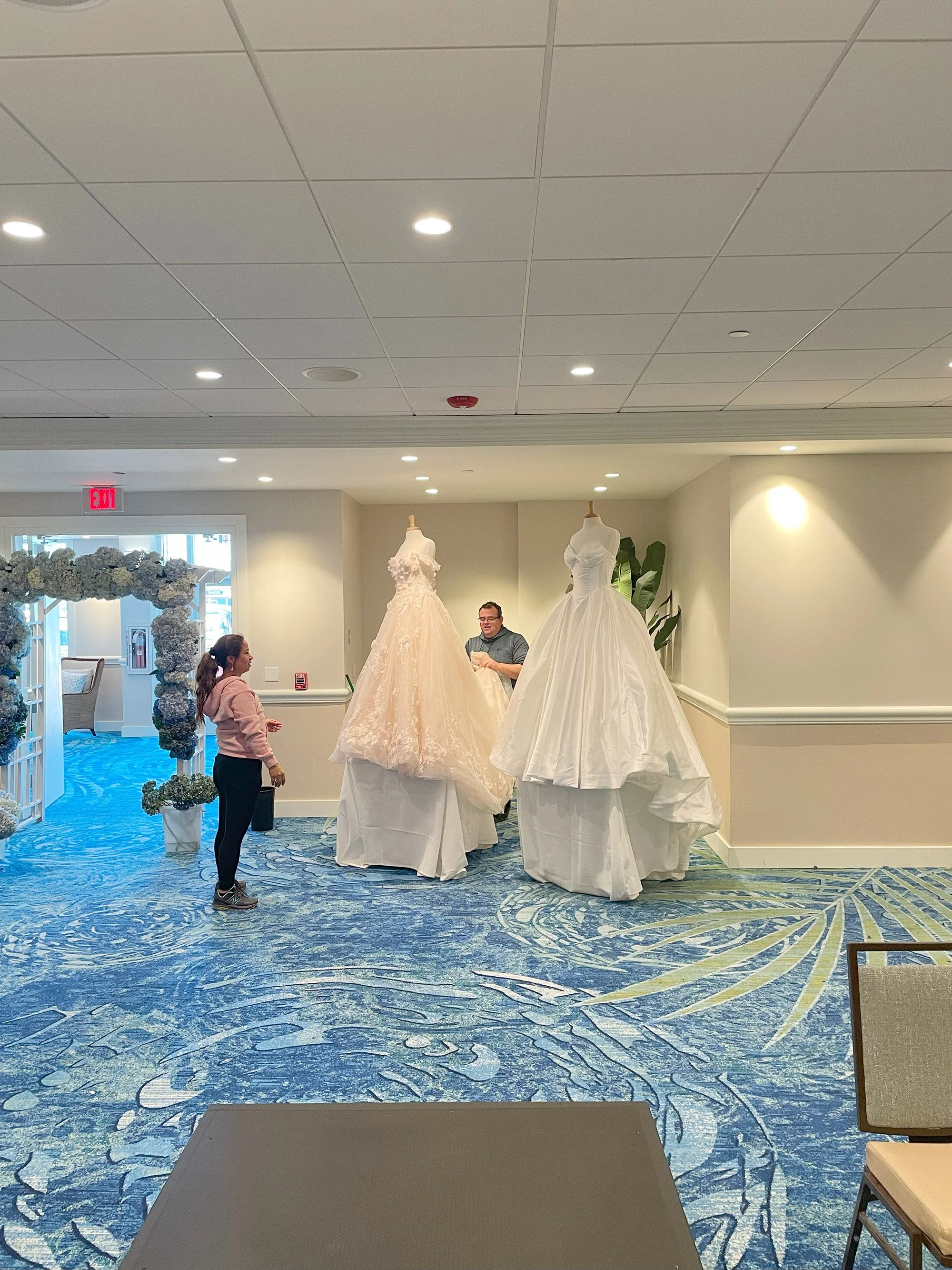 A woman and a man are in a bridal shop with two wedding dresses on mannequins, one on the left and one on the right. The woman is looking at the dresses, and the man appears to be preparing or adjusting them. There is a decorative arch made of flower