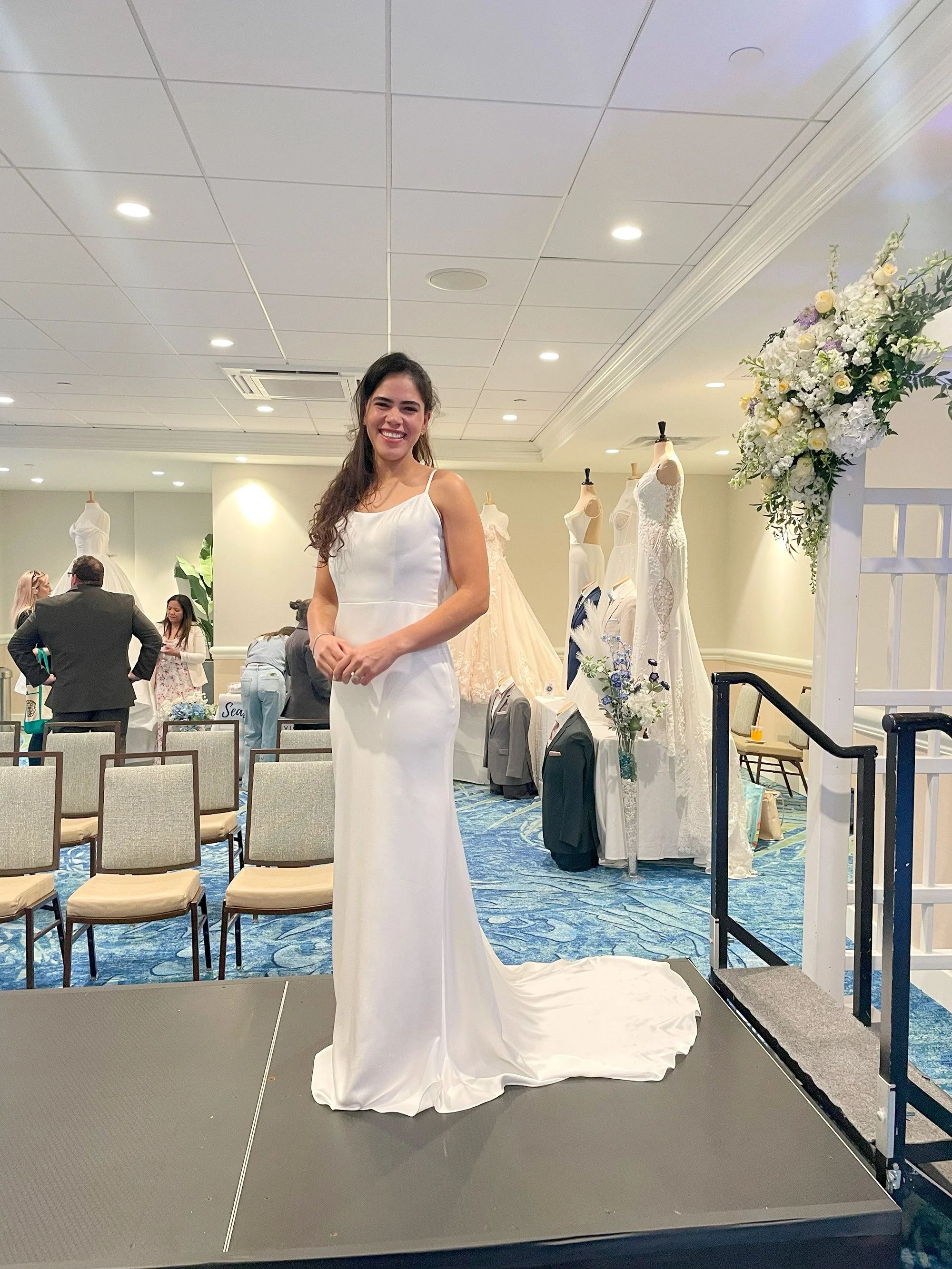 Young woman in a white wedding dress standing on a platform at a bridal shop, surrounded by wedding dresses and mannequins in the background.