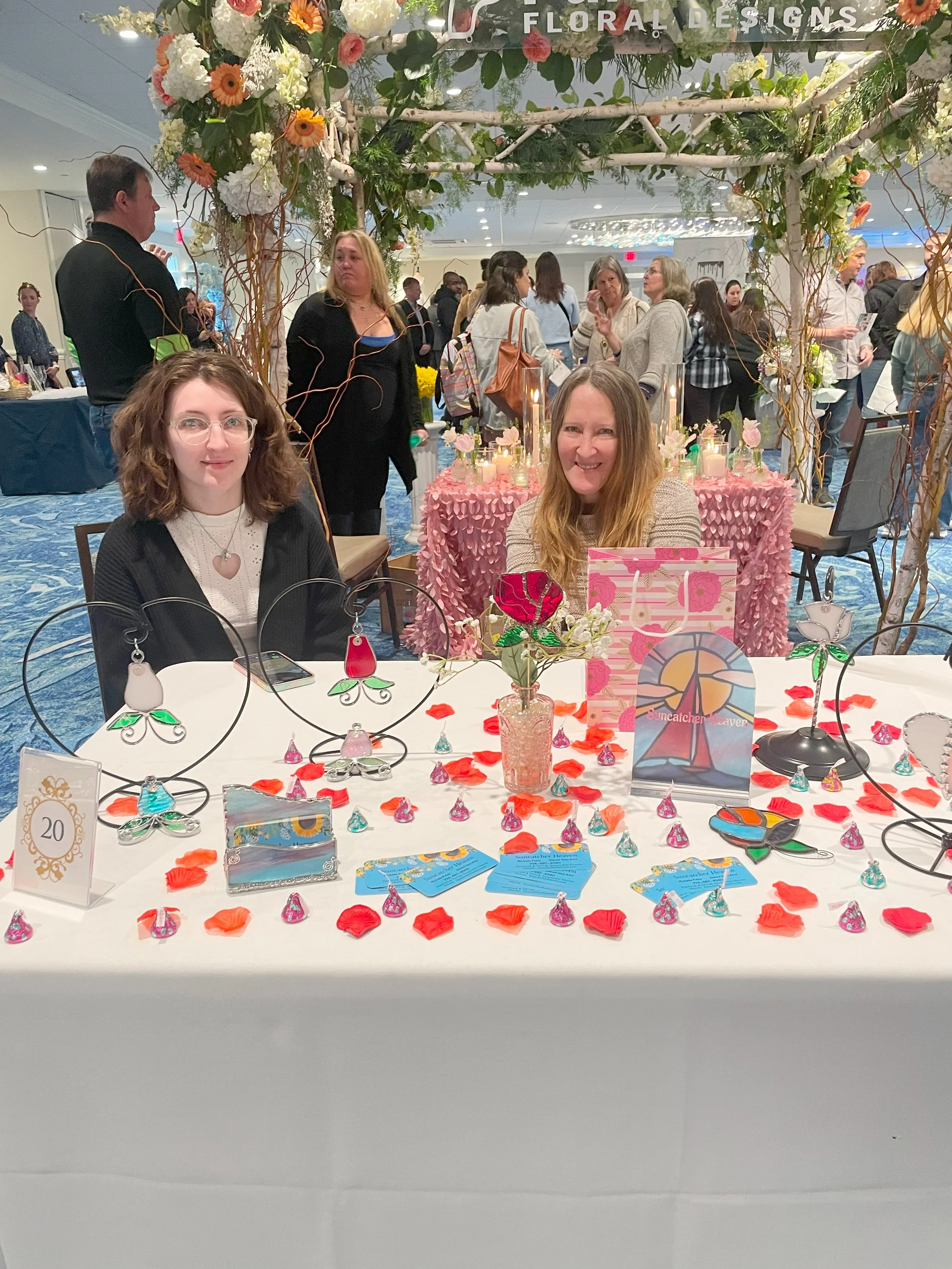 Two women sit at a decorated table with pink flower-themed jewelry and decorations, at a floral design event. A crowd is visible in the background with people mingling and browsing displays.