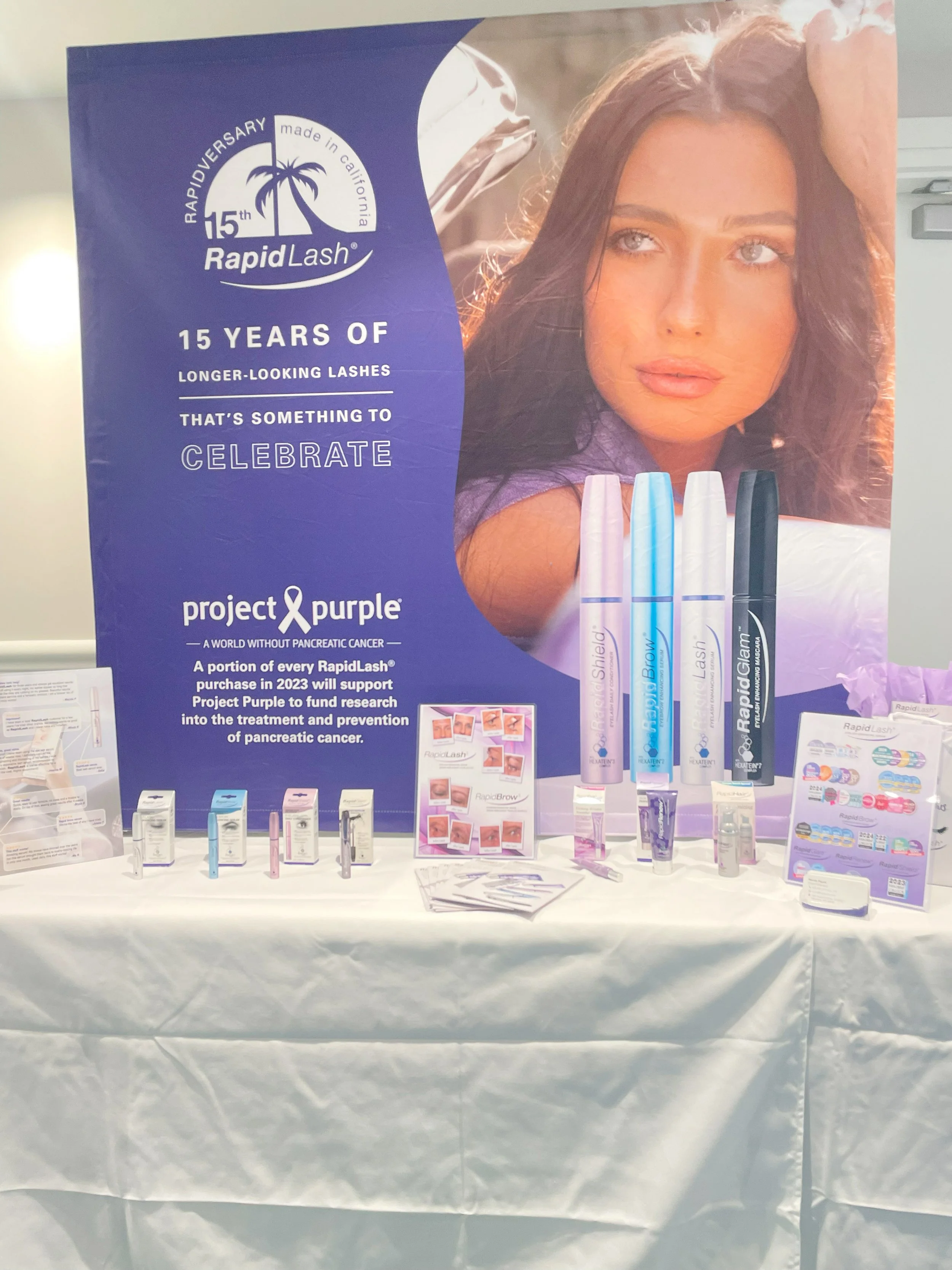 Display table of RapidLash beauty products with a purple promotional banner celebrating 15 years of longer-looking eyelashes and supporting Project Purple against pancreatic cancer, featuring a woman with long brown hair and a large advertisement wit