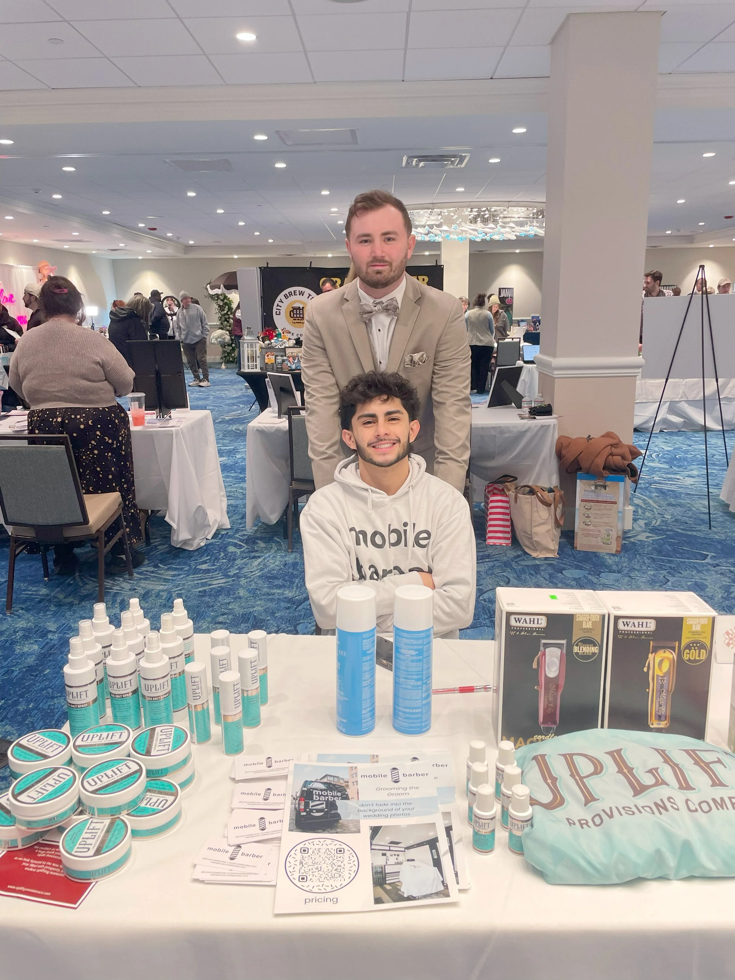 Two men at a promotional table, one sitting and one standing behind, at a busy indoor event. The table displays products labeled "UPLIFT" and promotional materials.