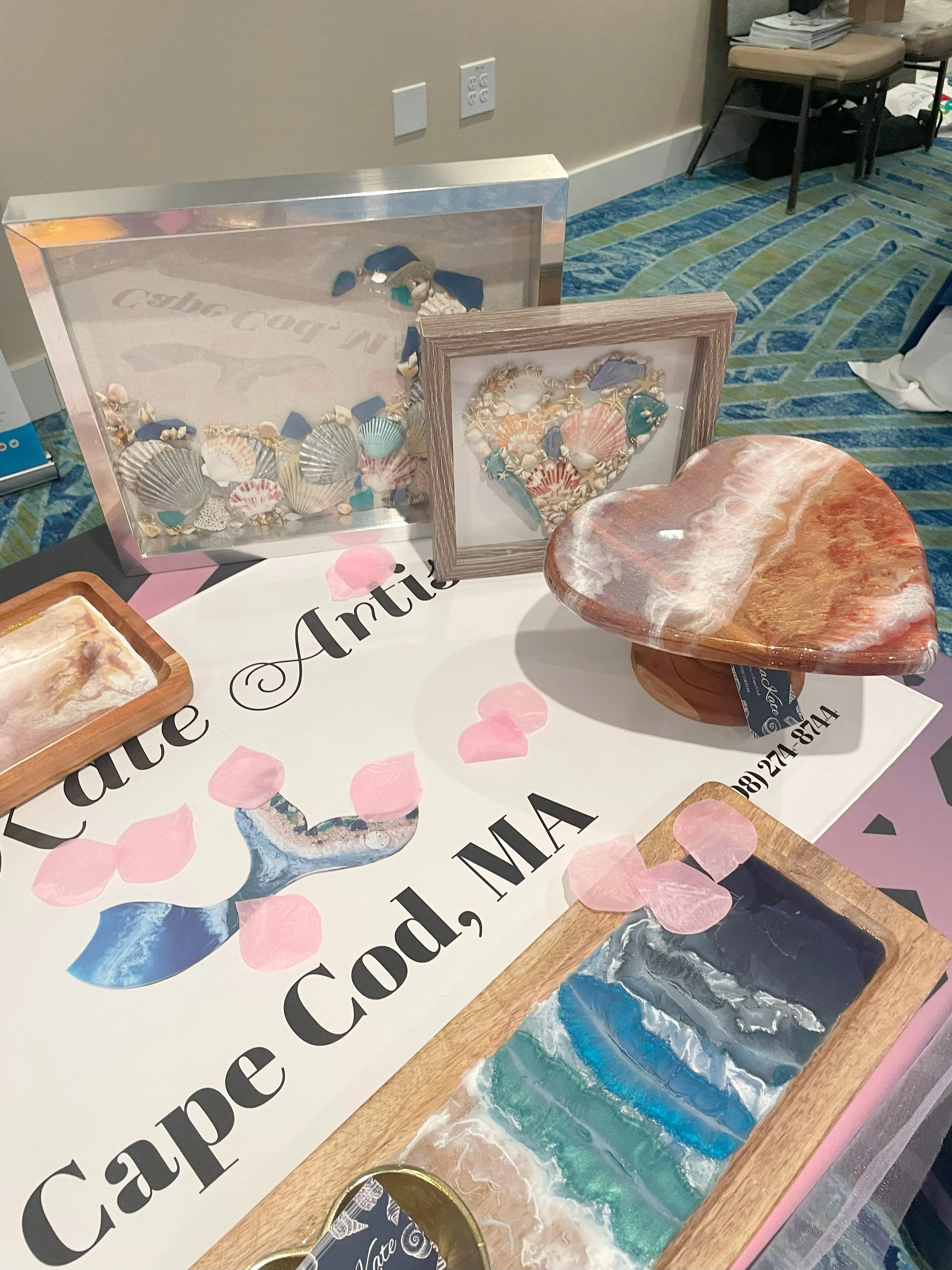 Display of seashell jewelry, framed seashell art, a heart-shaped shell art piece, and ocean-inspired resin art on a table with a "Cape Cod, MA" sign and pink flower petal decorations.