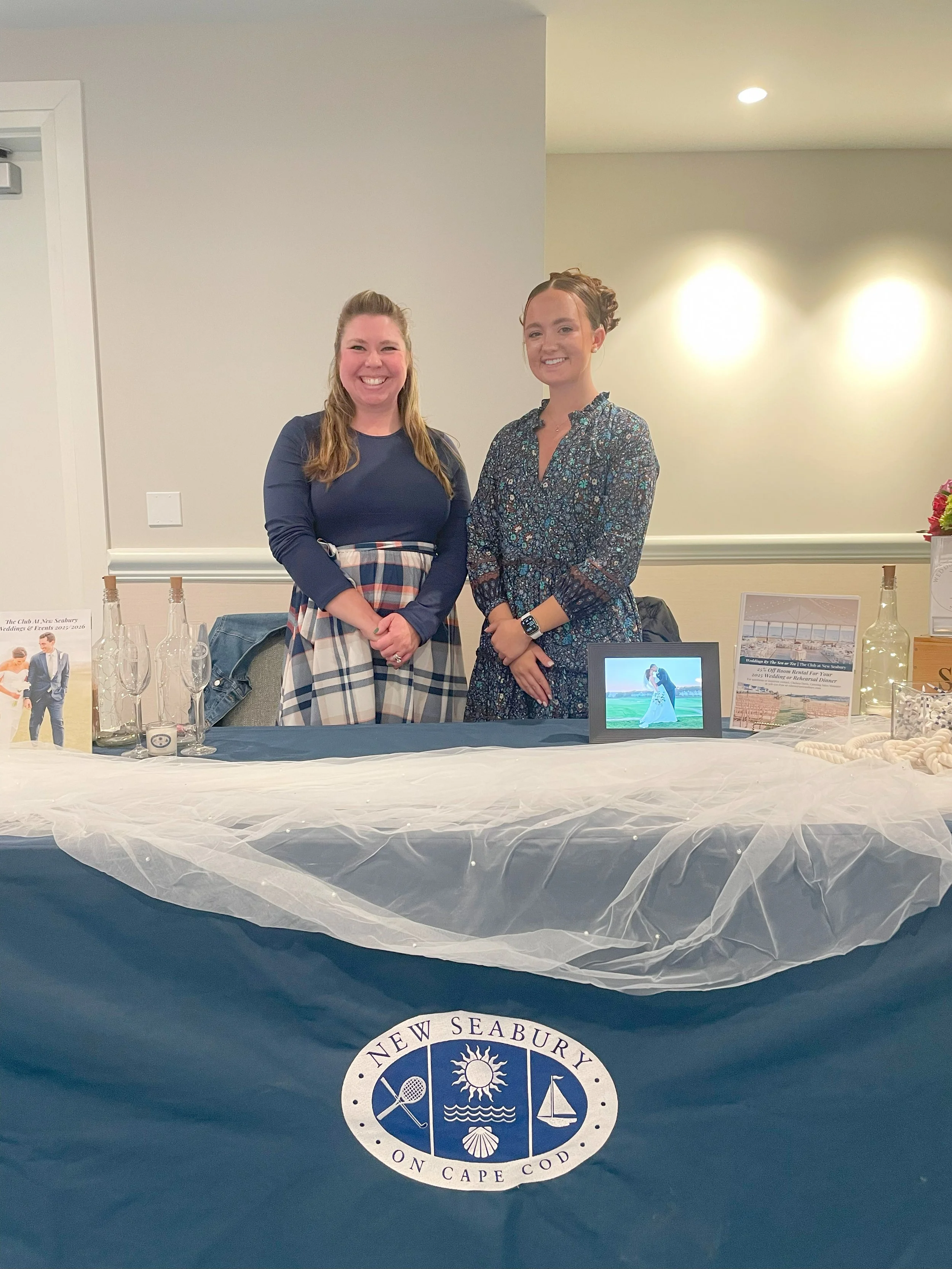 Two smiling women standing behind a display table with promotional materials and framed photos, at an event for New Seabury on Cape Cod.