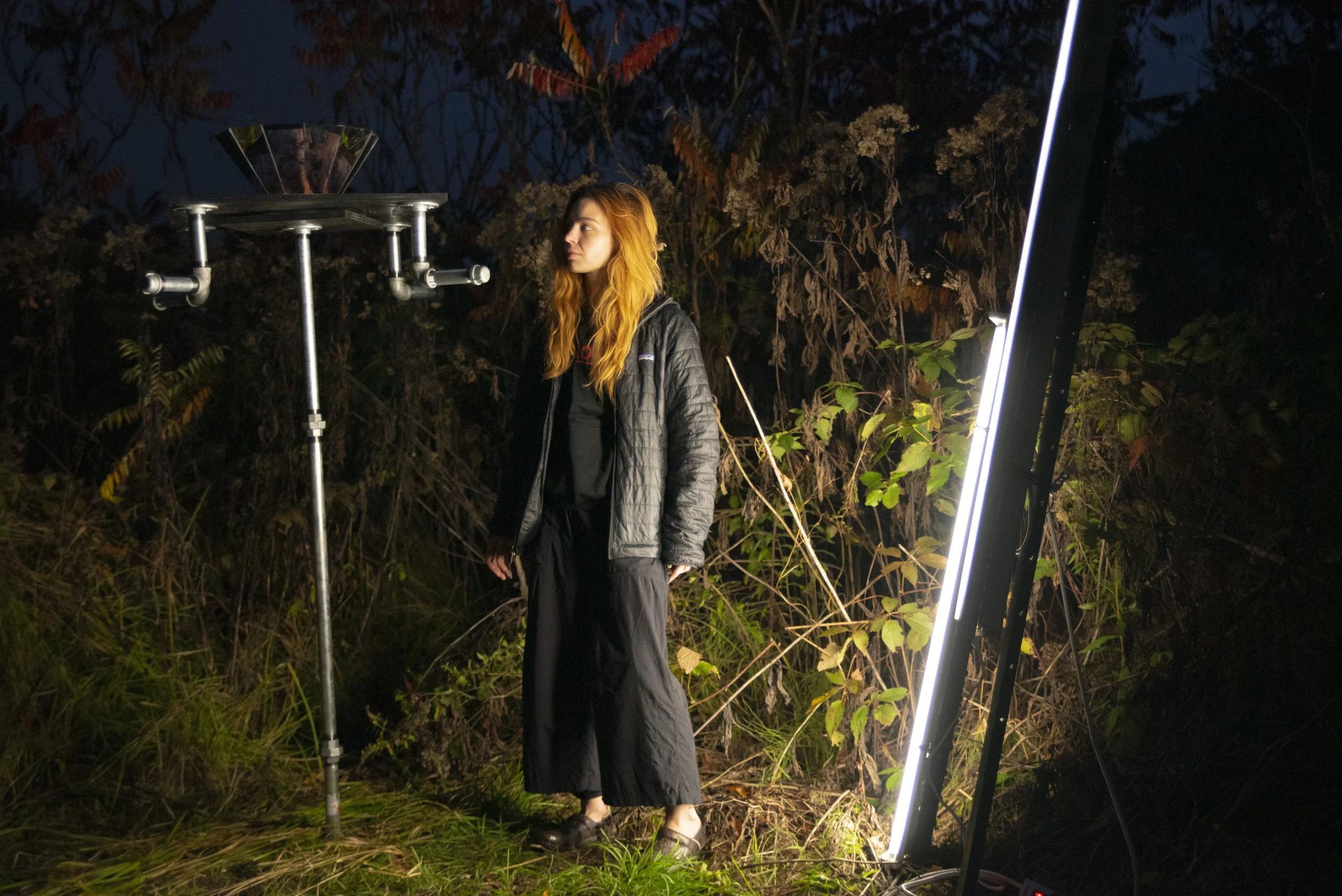 A woman with long orange hair standing outdoors at night surrounded by trees and bushes, illuminated by bright light from a large vertical LED panel on her right, with a metal pole with a small shelf and a reflective object on top to her left.