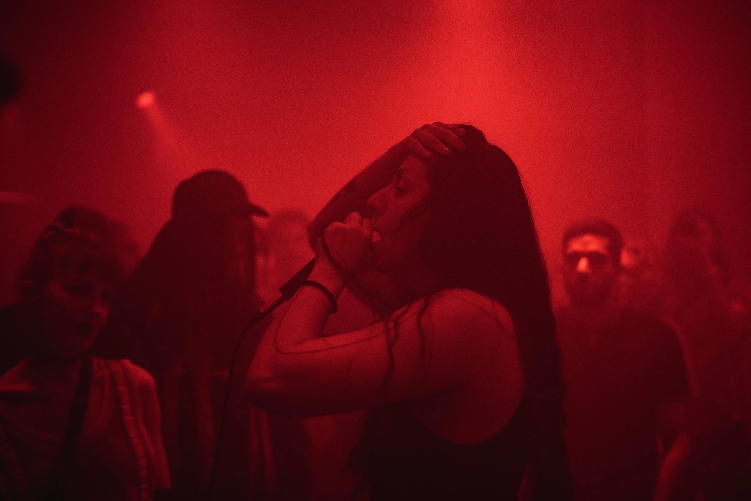 A woman with long dark hair singing into a microphone in a dimly lit, red-hued club or concert venue, surrounded by a few people.