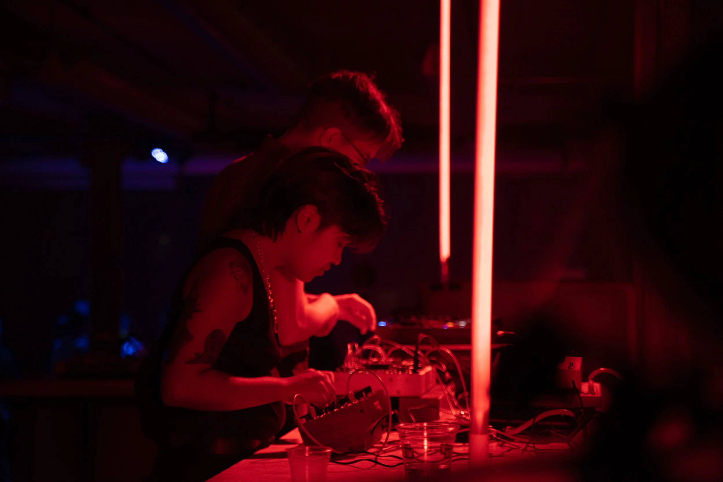 Two musicians perform at a dark, red-lit venue, one with tattoos and short hair, using electronic music equipment.