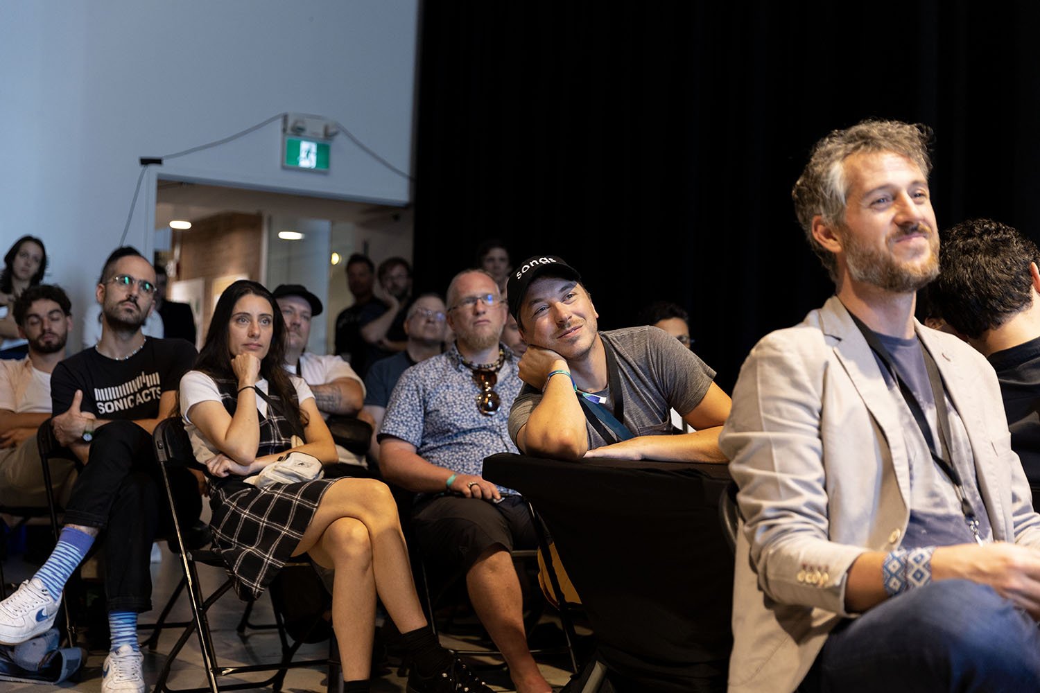 Audience seated and watching a presentation in a dimly lit room with black curtains.