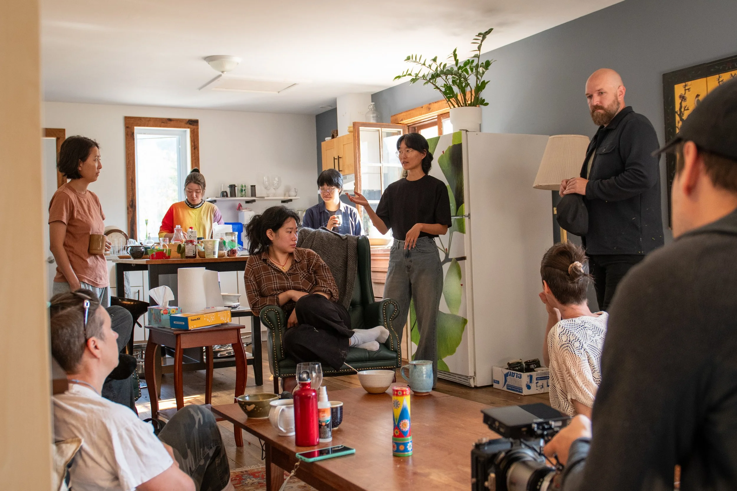 A group of people gathered in a cozy living space, with a woman standing and speaking to the group, while others sit and stand around listening. The room has a kitchen in the background and a variety of objects on the tables.