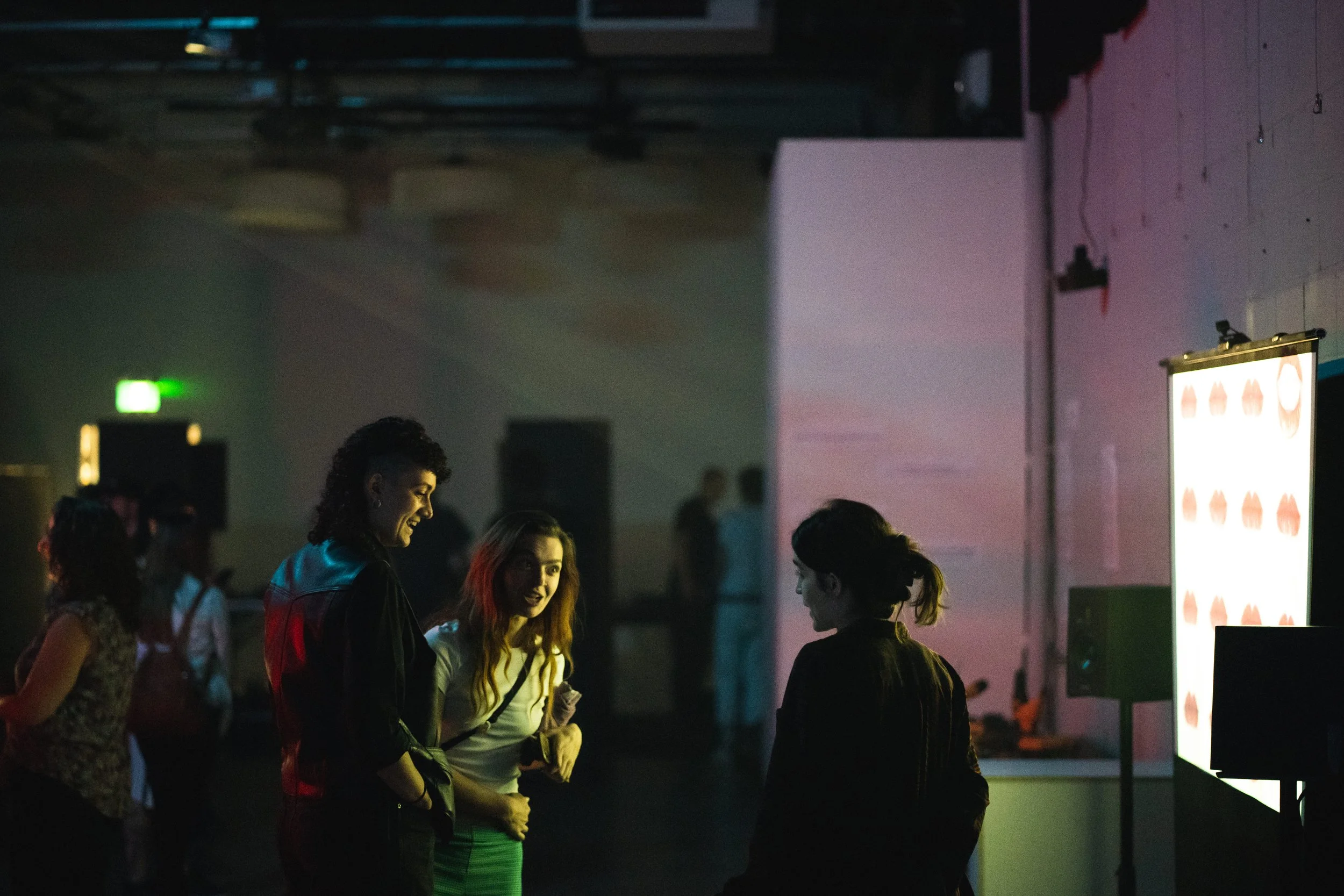 Three young women talking and smiling at an indoor event, with a large screen displaying a logo or pattern on the wall to their right.