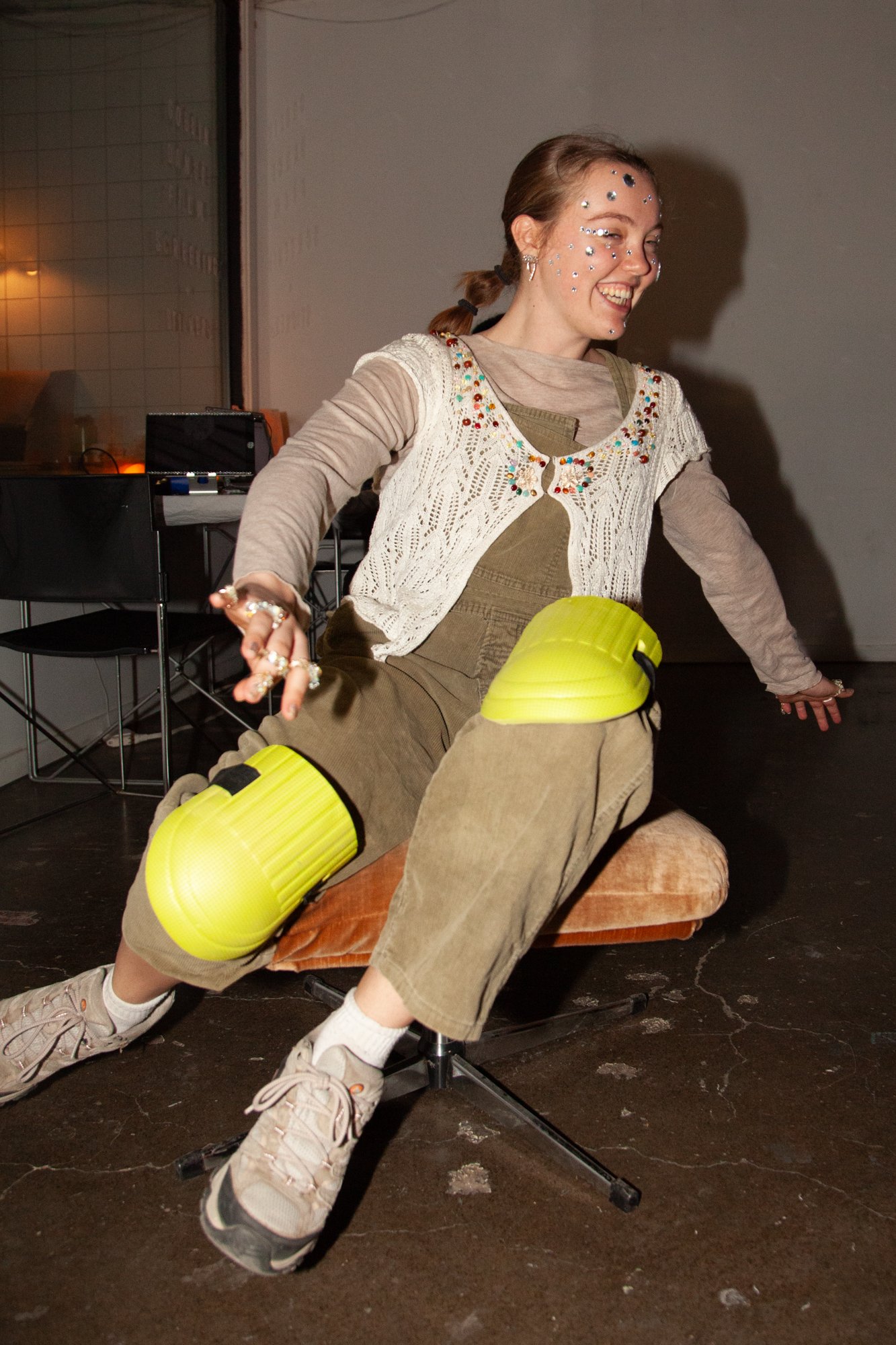 A young woman sitting on a chair with her legs extended, laughing joyfully. She has face gems, is wearing a beige long-sleeve shirt with a cream crocheted vest, and olive pants with yellow knee pads. The setting appears to be indoors with a dark floo