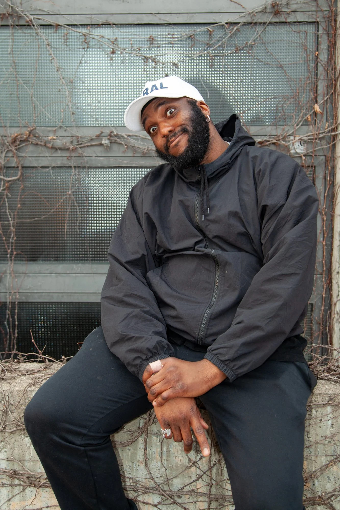 Adrian sitting outdoors, wearing a white cap, black jacket, and black pants. Background features a metal screen covered with dried vines.