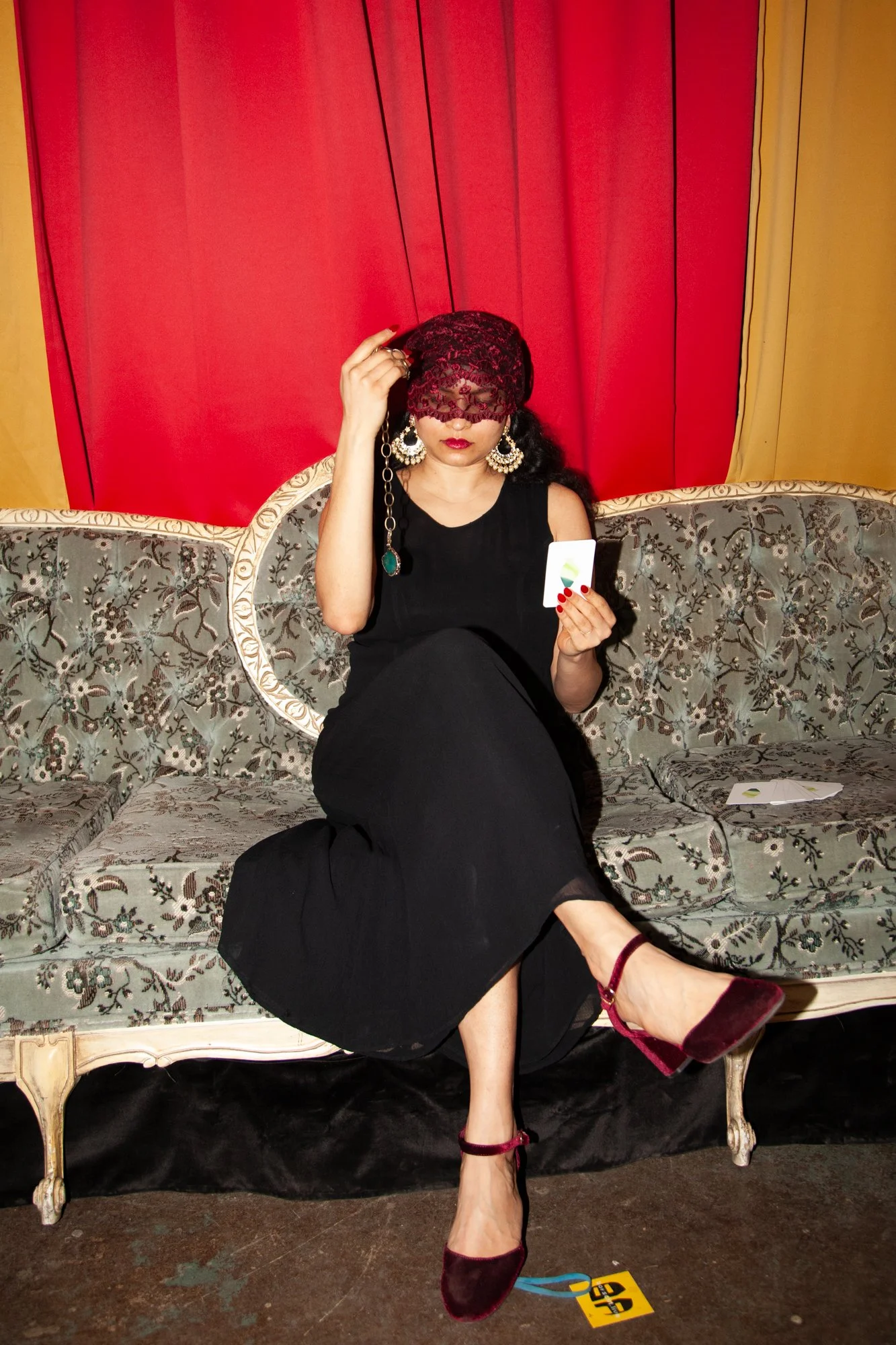 A woman in a black dress and burgundy high heels sitting on a vintage sofa. She wears large earrings and a lacy burgundy eye mask, holds a card in one hand, and adjusts her mask with the other. Red curtains are behind her.
