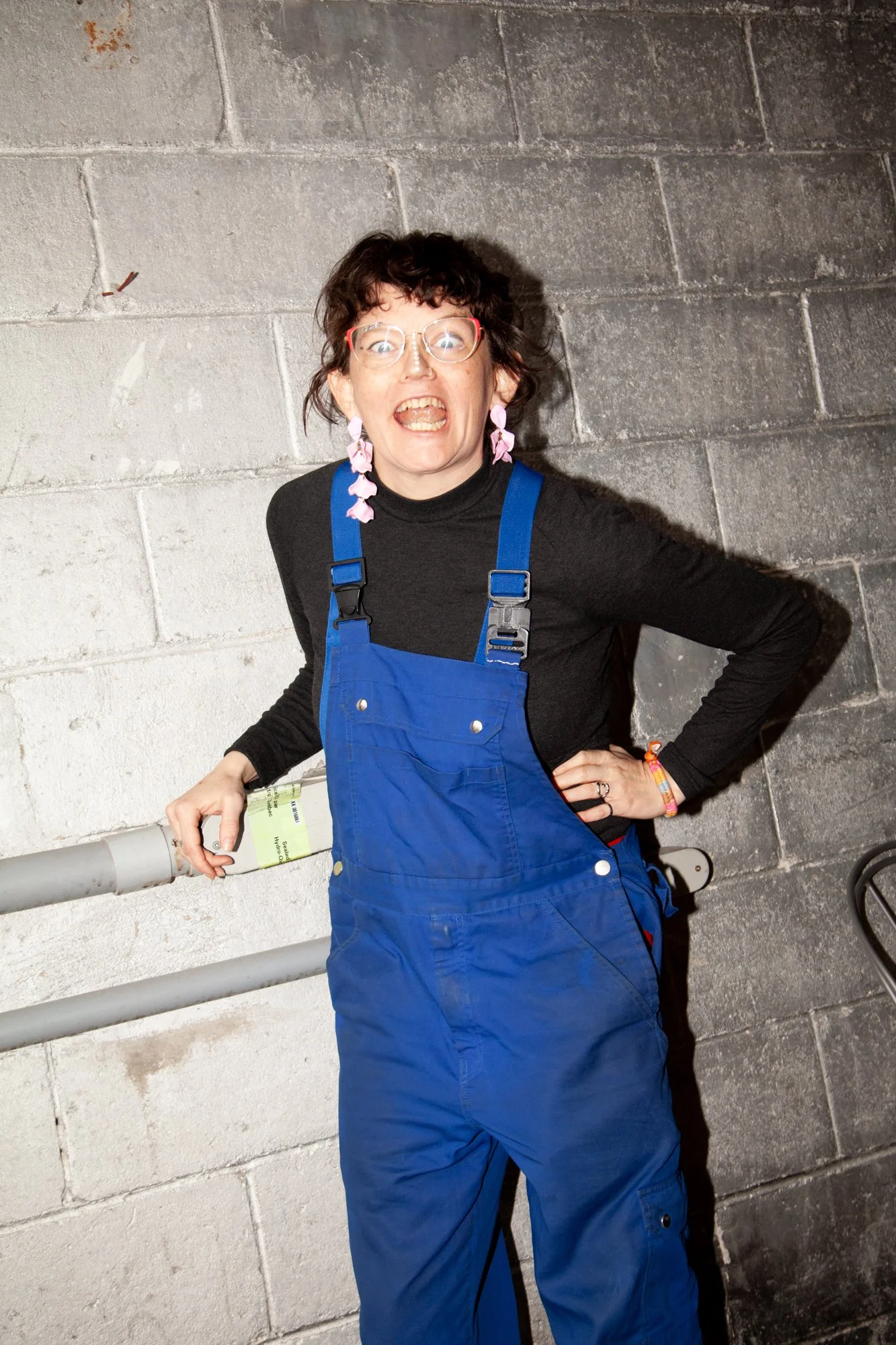 A woman with curly dark hair wearing glasses, pink earrings, a black turtleneck, and blue overalls, standing against a gray concrete wall and holding a book in her right hand.