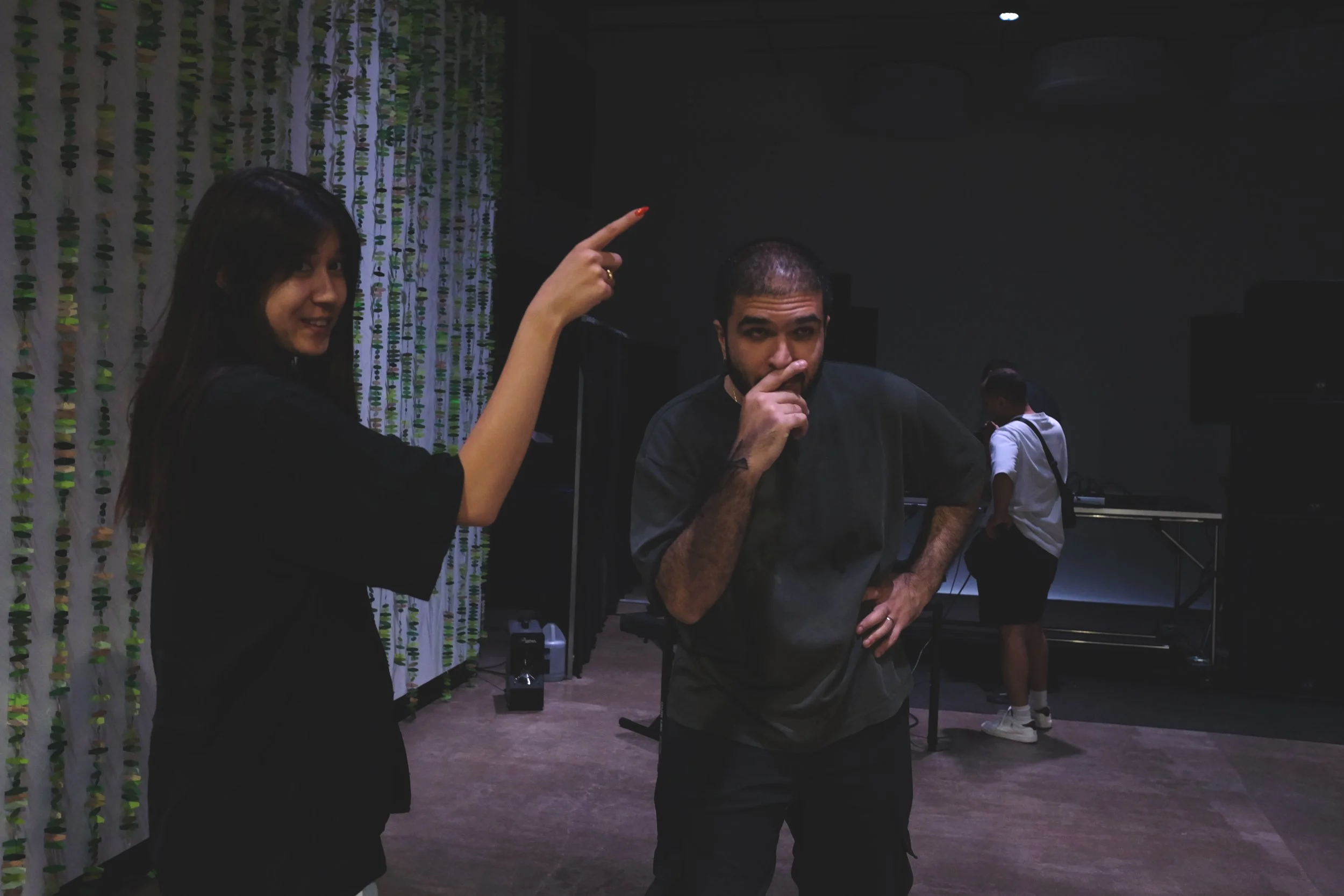 A woman with long dark hair pointing at a man with short hair and a beard, who is thinking with his finger on his lips in a dark room with a patterned curtain, a person in the background working with electronic equipment, and another person facing aw