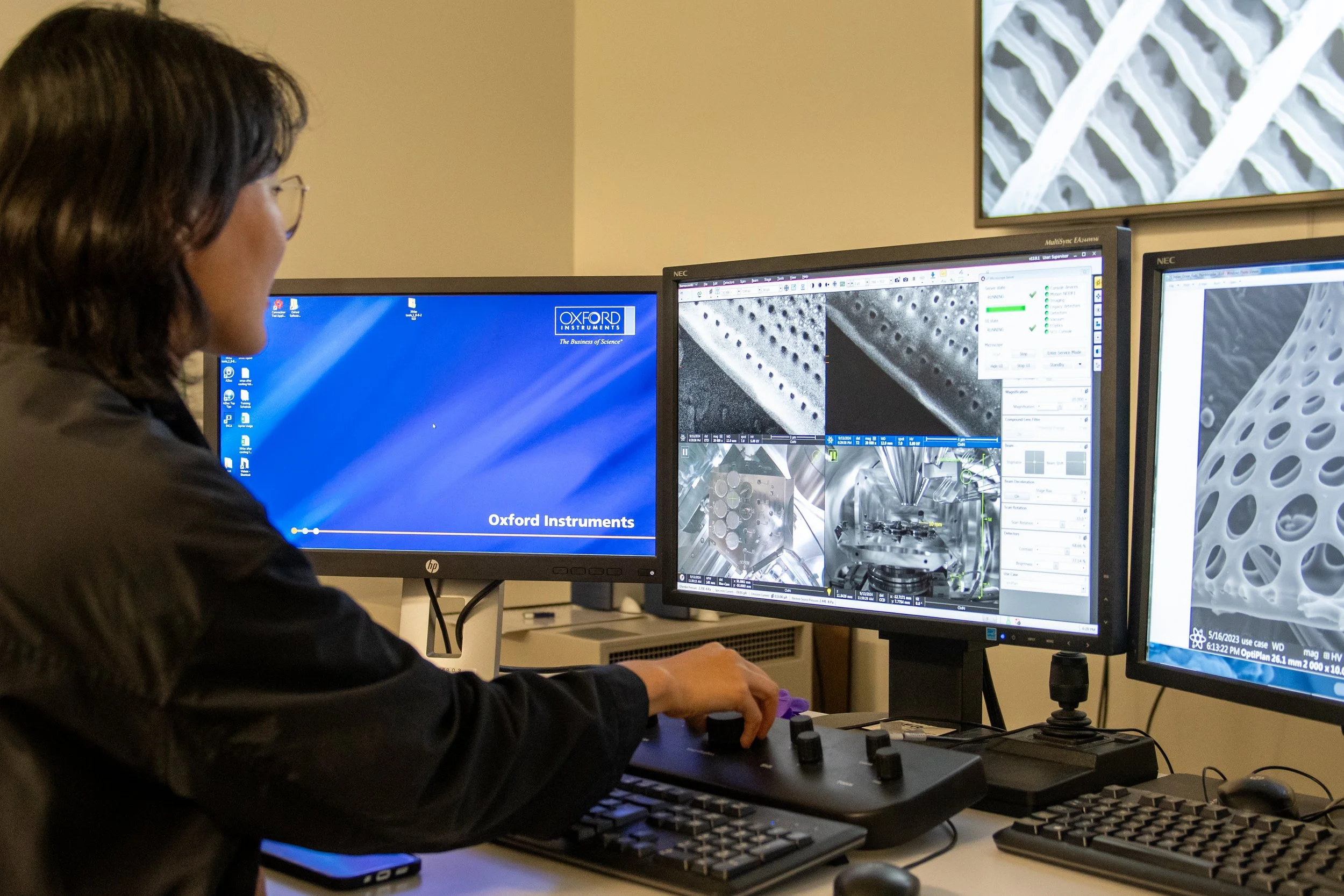 A researcher working at a desk with three computer monitors, analyzing microscopic images of a material with a pattern of holes, in a laboratory setting.