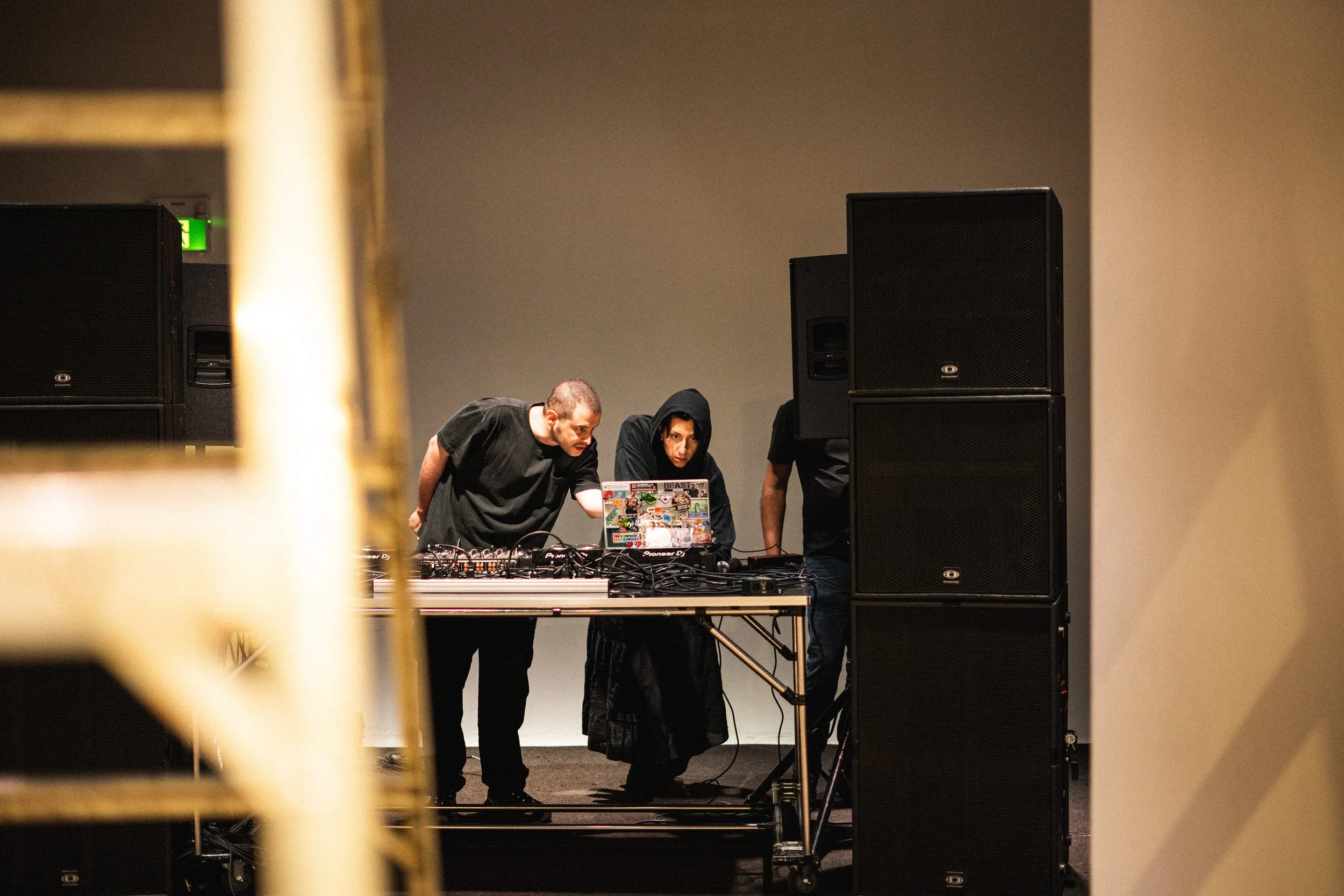 Three people working at a DJ booth, two of them dressed in black, one with a hoodie, with large speakers on each side of them, viewed through a gap in a golden railing.