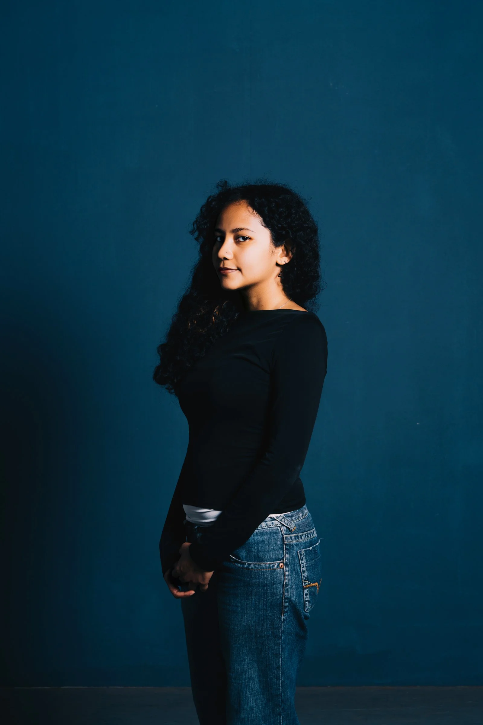 A young woman with curly dark hair standing against a plain dark blue wall, wearing a long-sleeve black top and blue jeans.