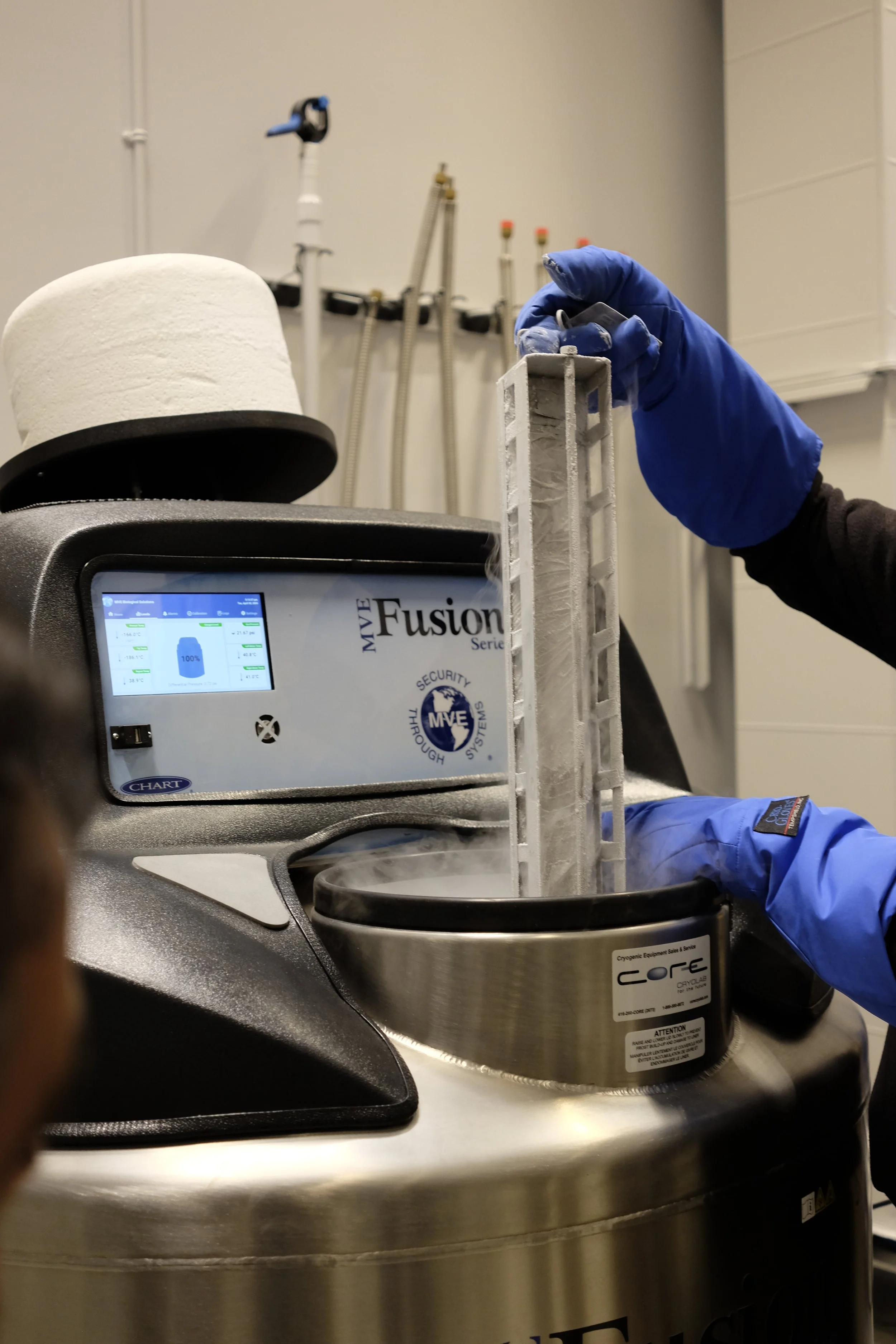 A scientist in blue gloves handling a white, textured sample inside a metal container connected to a CryoTherm system, with a bio-freeze device and laboratory equipment in the background.