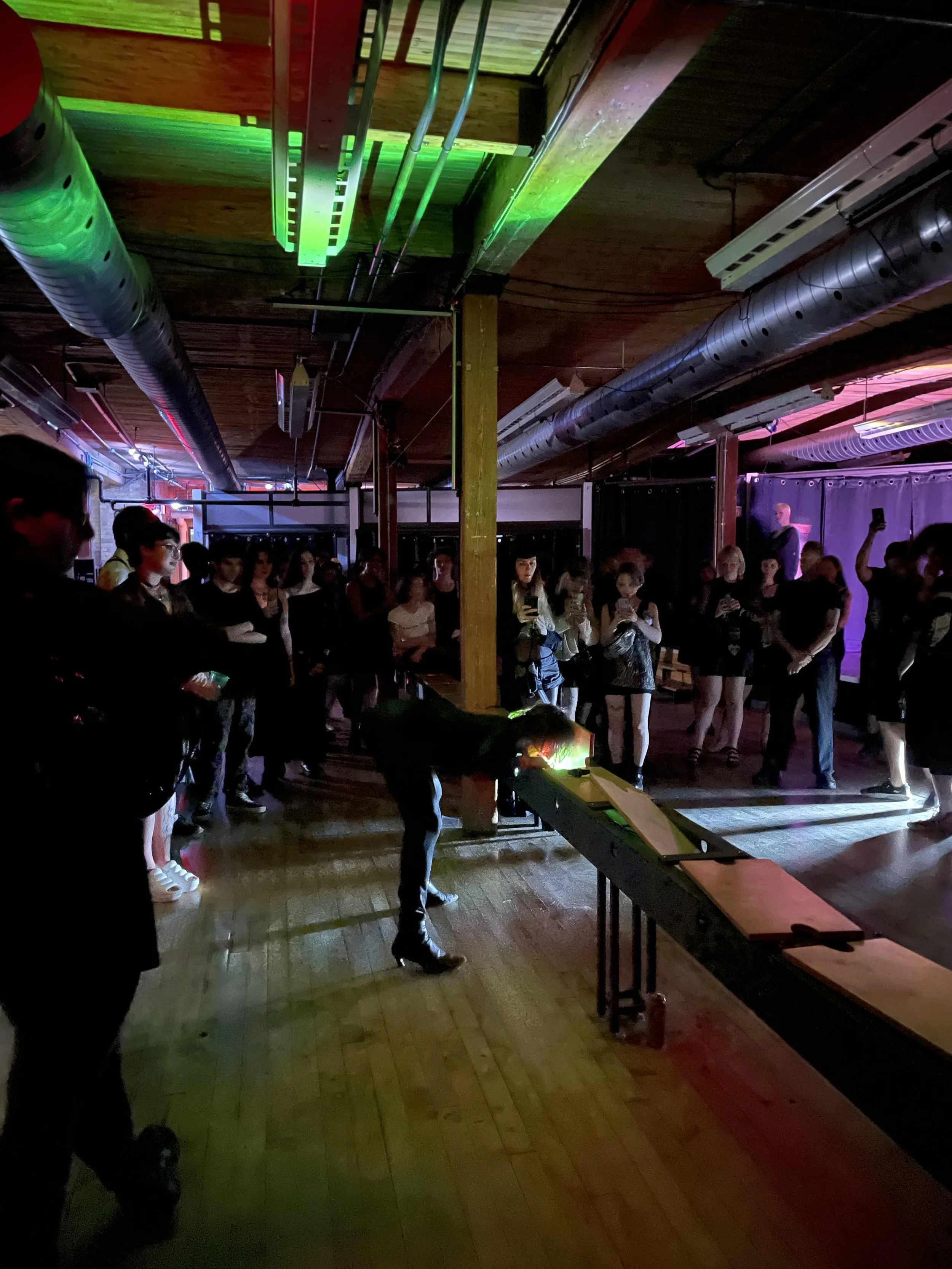 People gathered in a dimly lit industrial-style venue with pipe ceilings, watching a person in high heels perform on a table under colorful lighting.