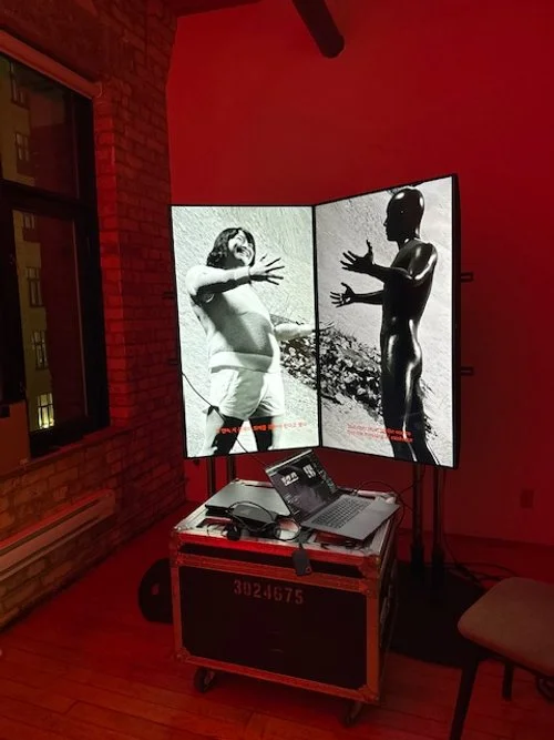 A digital art installation featuring two black-and-white photographs of a child and a puppet on adjacent screens, surrounded by a red-lit room with balcony windows.