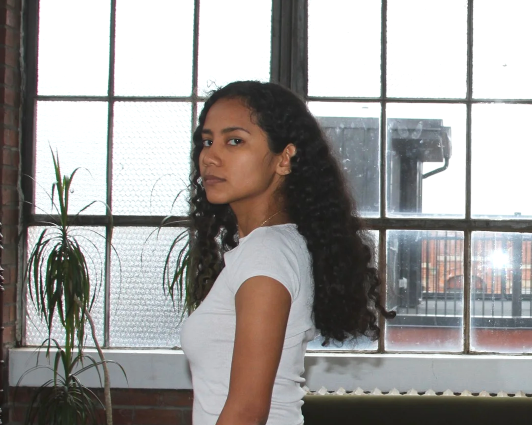 A young woman with long, curly black hair and medium skin tone standing indoors near a large window with metal framing. She is wearing a white t-shirt and looking at the camera with a neutral expression. There are indoor plants and brick walls in the background.