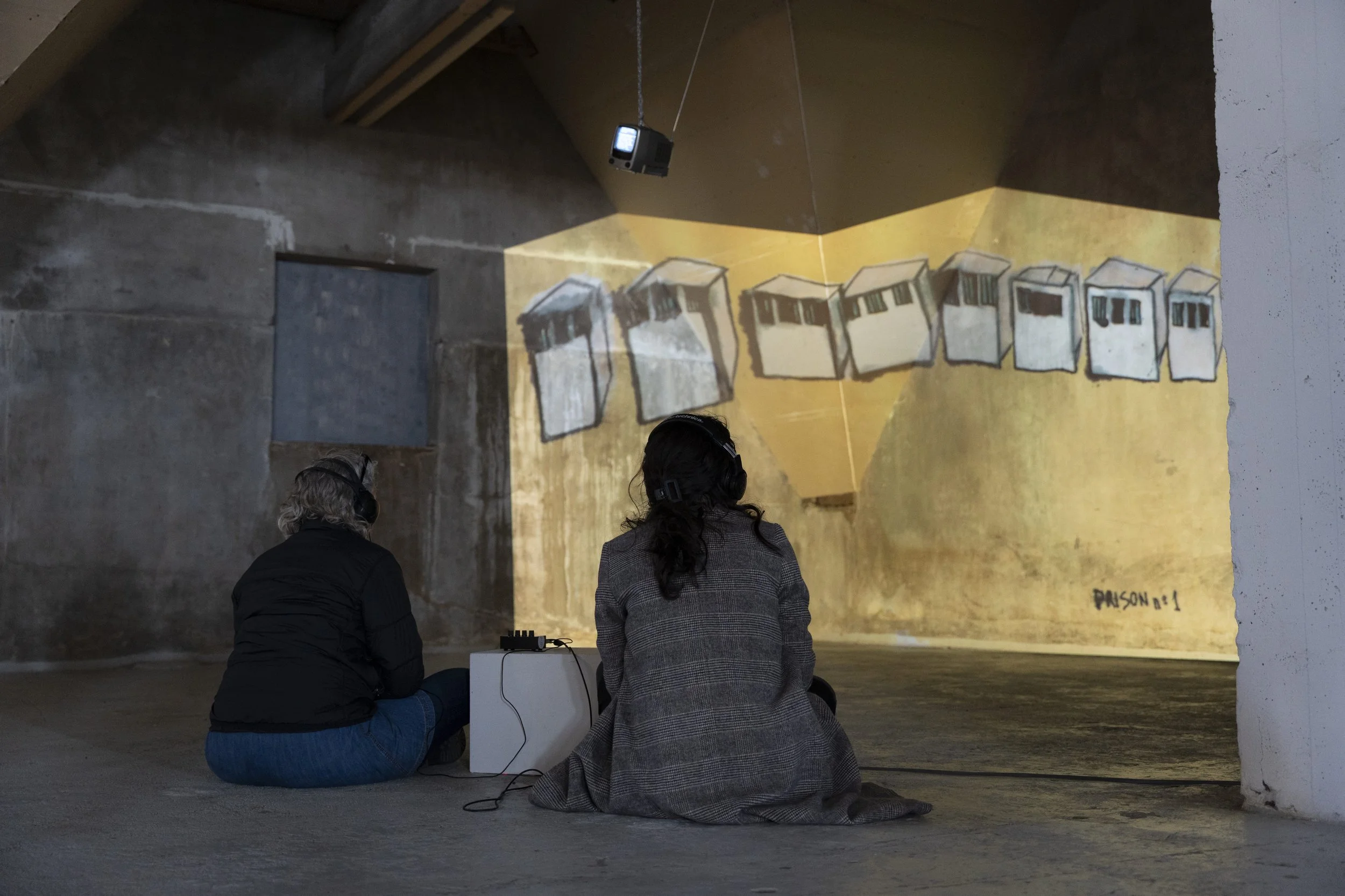 Two people sitting on the floor in a concrete room, watching a projection of abstract geometric shapes on the wall, with headphones and a small audio device.