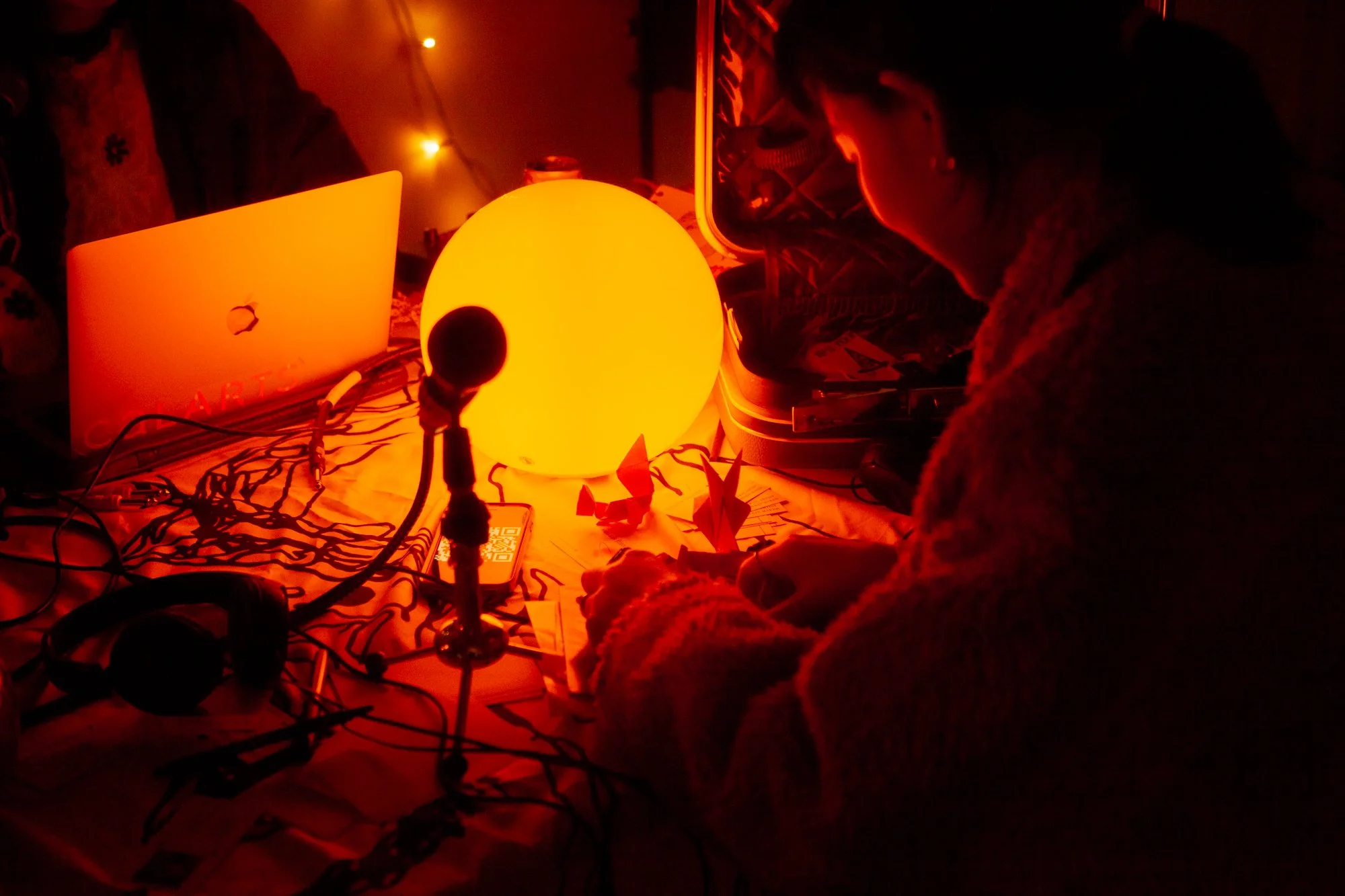 A person in a cozy sweater sitting at a cluttered table illuminated by a glowing orange orb lamp, with origami figures and a microphone in front. The scene has warm, red-orange lighting and a string of small lights in the background.