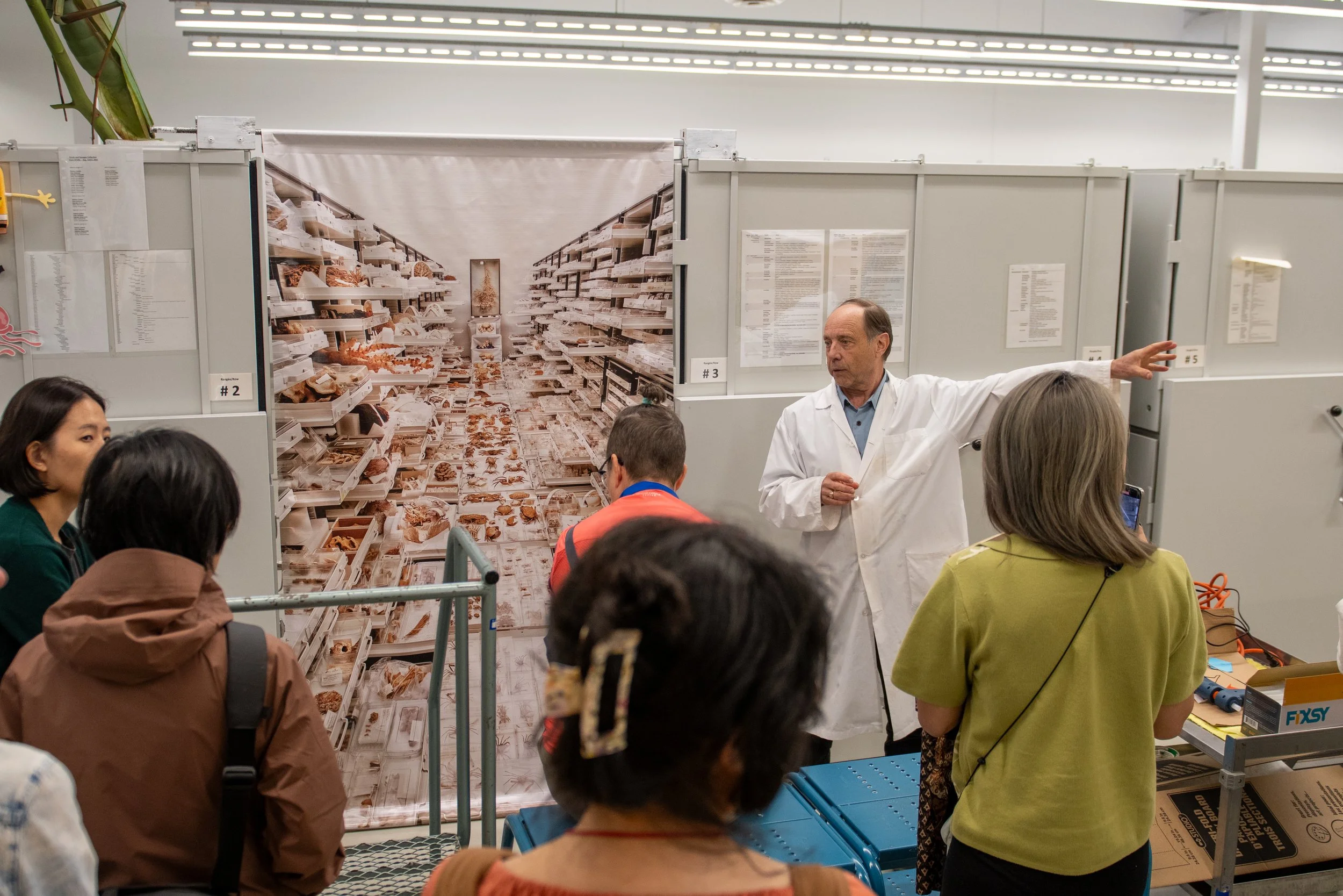 A man in a white lab coat giving a presentation to a group of people in a store aisle with shelves filled with snacks or baked goods and a giant photograph of a similar aisle in the background.