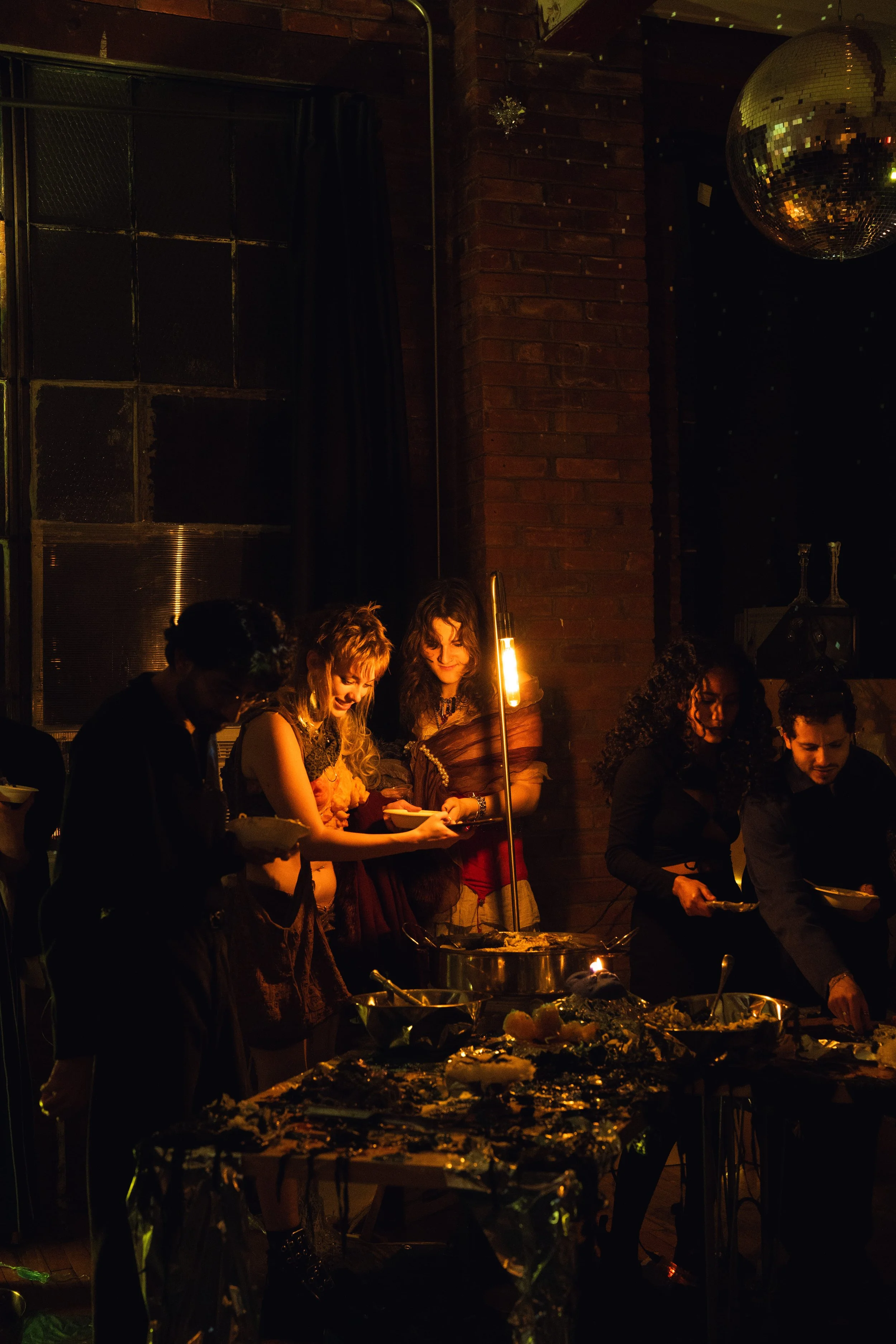 A group of people at a dinner party in a dimly lit space, serving food from a table, with a disco ball hanging from the ceiling.
