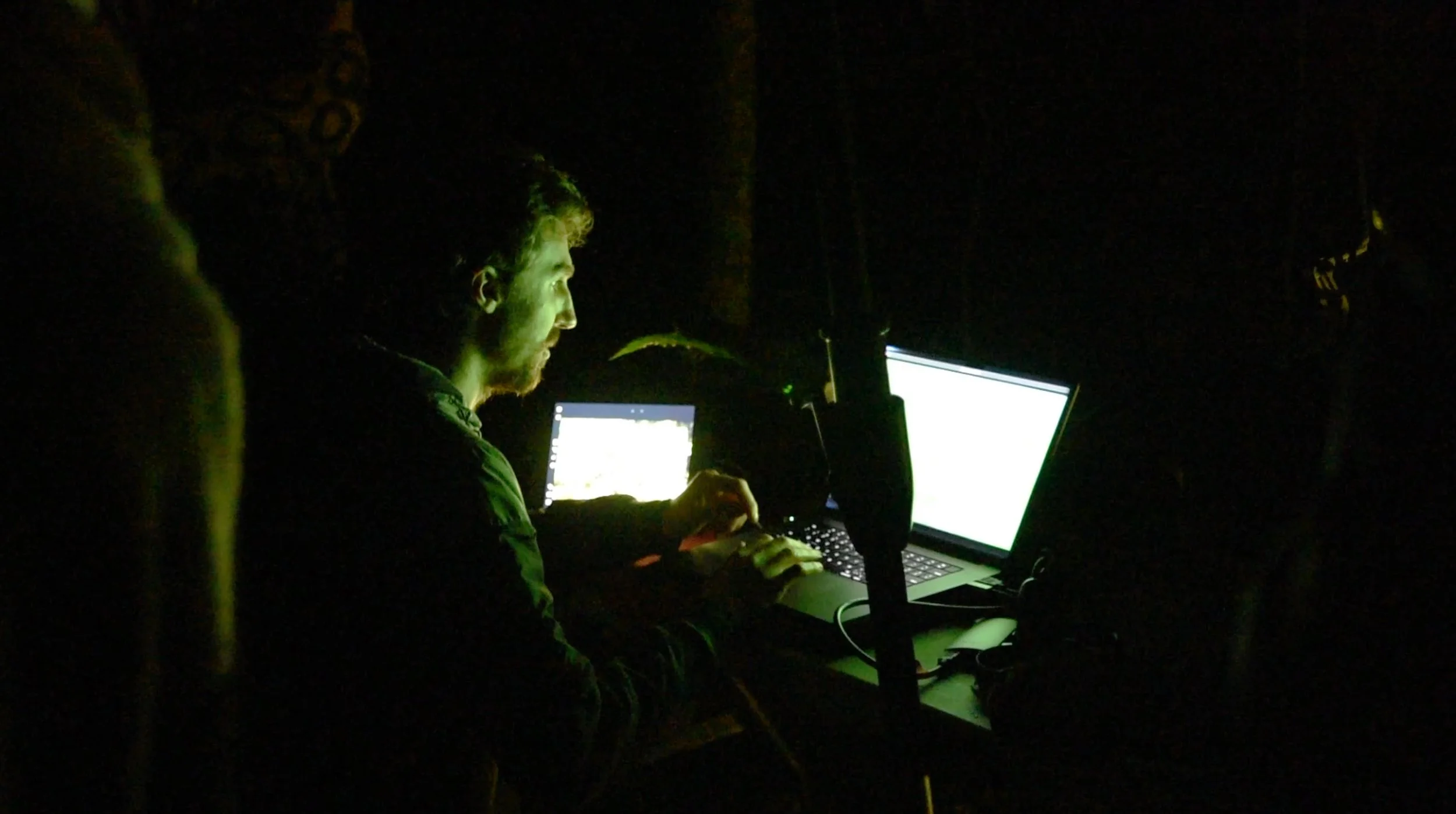 A person working on a laptop in a dark environment, illuminated by the screens, with a microphone and a smaller electronic device nearby.