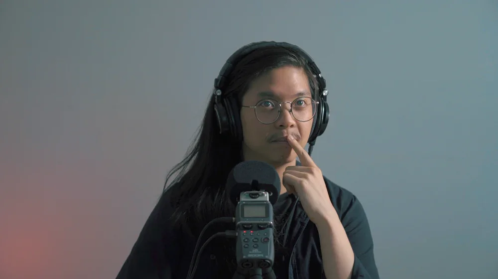 A person with long dark hair, glasses, and headphones, holding a finger to his nose, sitting in front of a microphone.